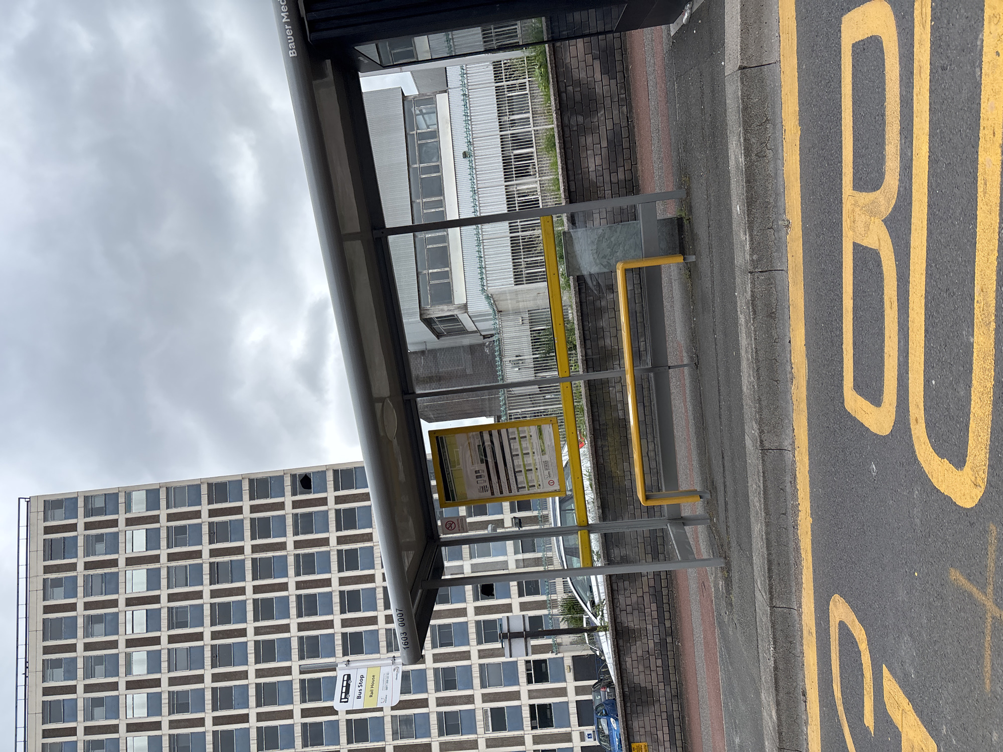 The image shows a bus stop shelter with a yellow bench and timetable. The bus stop is labeled "Bus Stop Rail House". In the background, there is a tall, multi-story building with many windows. The bus stop is located on a street with yellow markings on the road, indicating a no-parking zone. The sky is cloudy. The bus stop appears to be located on the outskirts of Crewe, United Kingdom.