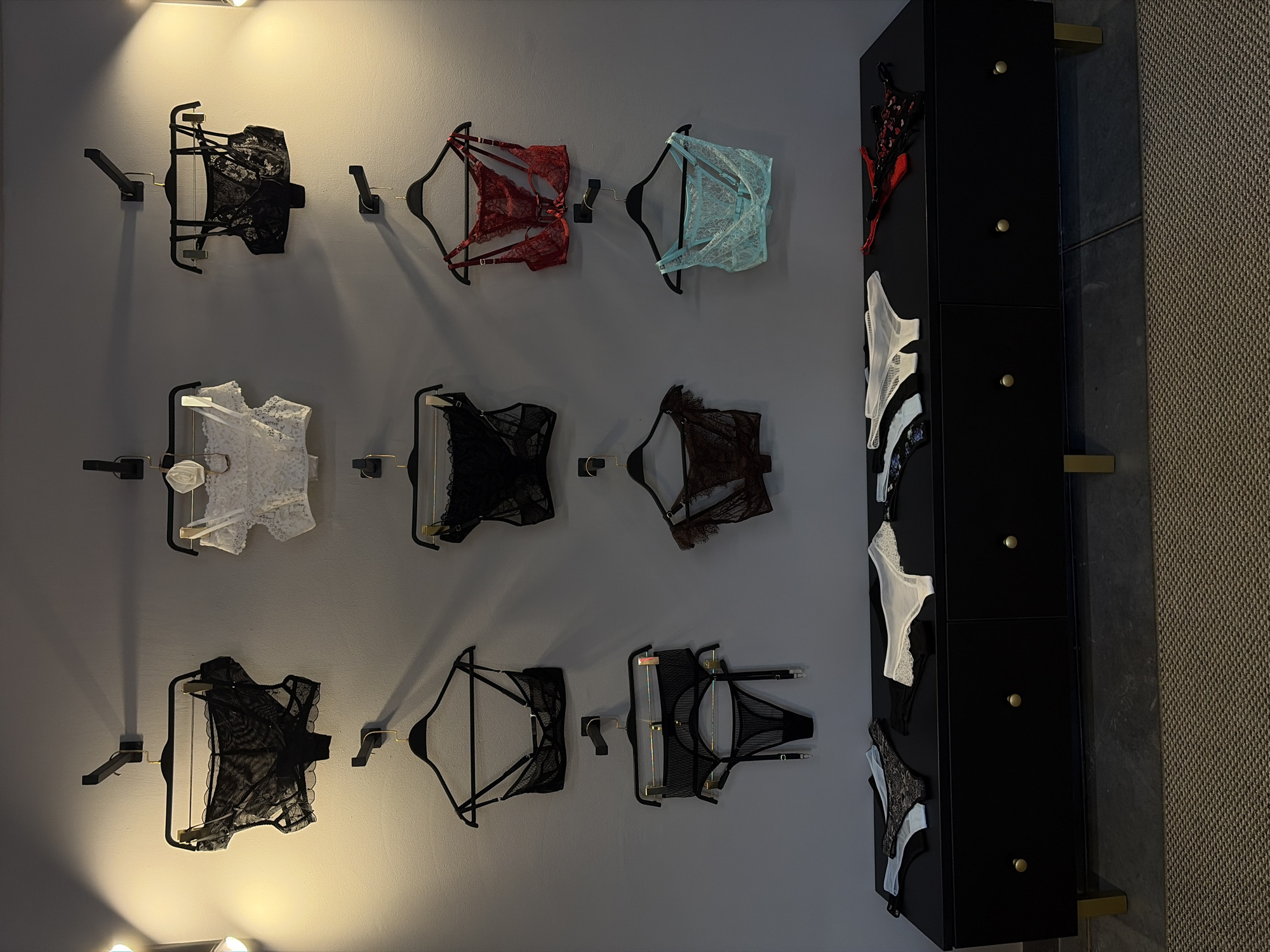 The image depicts a wall display of lingerie items in what appears to be a retail or boudoir setting. Several bras and matching briefs or garter sets are hung on black hangers attached to wall hooks. The items are arranged in three rows of three. The top row features a black lace bra, a white lace bralette with floral embellishments, and a black lace bra with thin straps. The middle row displays a black sheer bra, a red lace bra and thong set, and a mint green lace bralette. The bottom row shows a black bra and matching briefs with garter straps, a brown lace set, and a teal lace bralette. Below the wall display, a black dresser with gold hardware holds several pairs of folded panties. The lighting in the scene is dramatic, with spotlights casting shadows on the wall. The background is a plain gray wall, and the floor is covered with a textured rug and dark flooring. There are no people visible in the image. The setting is a room with a clean, modern aesthetic. The location is specified as Chaikovsky, Russia.