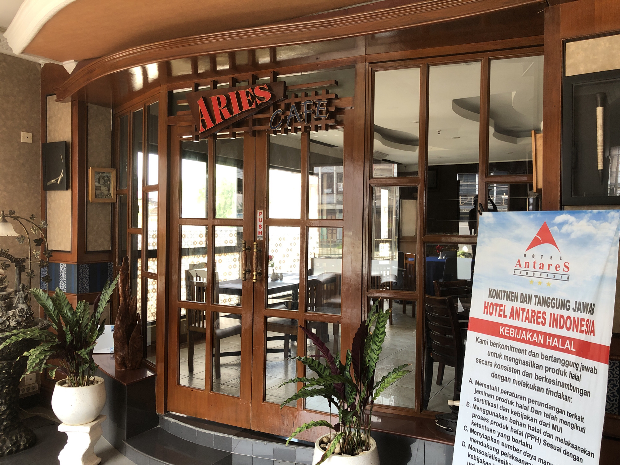 Here is a detailed description of the image:
**Scene:** The image shows the entrance to "Aries Cafe", which is part of the Hotel Antares Indonesia in Medan City, Indonesia.
**Entrance:** The cafe's entrance has a wooden frame and glass panels, and double doors with gold handles and "push" labels. A sign above the doors reads "ARIES CAFE" in red (Aries) and blue (Cafe).
**Decor:** Potted plants (fern-like) sit on either side of the entrance. There's decorative wood art (or a natural wooden element) to the left. Through the glass, tables and chairs can be seen inside the cafe.
**Hotel Signage:** To the right of the entrance, there's a sign for Hotel Antares Indonesia, declaring their "Kebijakan Halal" (Halal Policy). The sign outlines their commitment to producing halal products consistently.
**Ambience:** The overall feel is somewhat dated, with the brown wooden decor. The scene looks to be during the daytime, with natural light coming through the windows.