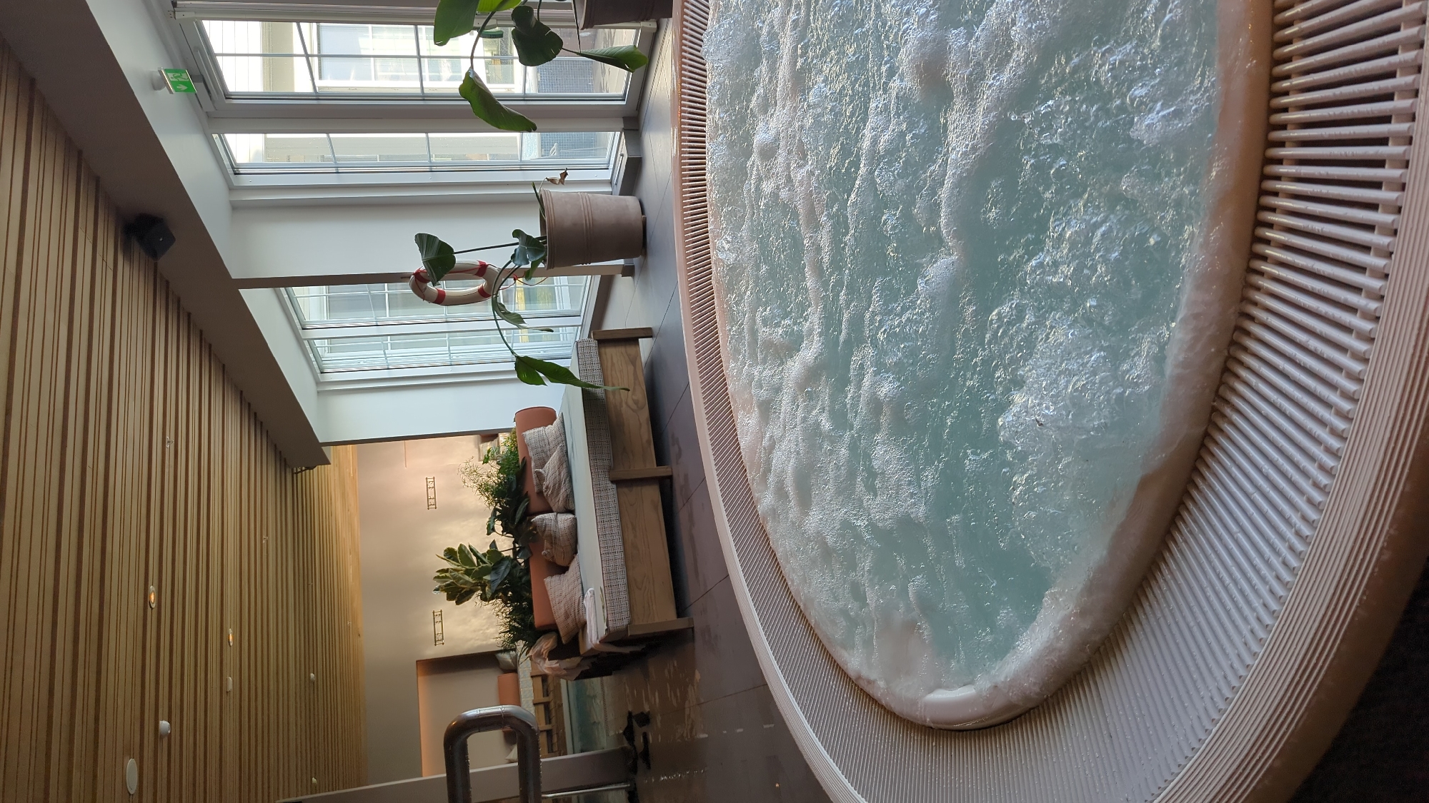 The image shows an indoor hot tub with bubbling blue water in the foreground. The hot tub has a white ribbed edge. In the midground, a wooden daybed with cushions and pillows is visible, positioned near a large window. Several potted plants are placed around the daybed and near the window. Behind the daybed, there appears to be another pool area and lounge chairs. The ceiling is made of wooden slats. The overall environment suggests a spa or wellness area. The image is located in Larkollen, Norway.