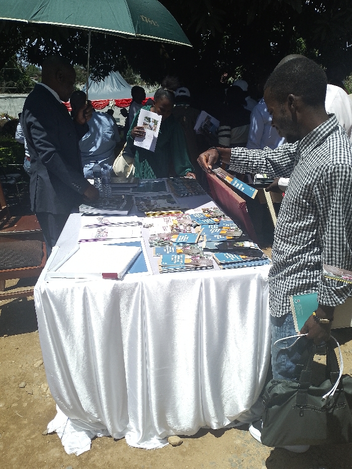 The image shows a table covered in white fabric, located outdoors on a dirt ground. The table is covered in a variety of pamphlets and brochures. A man in a plaid shirt stands by the table, reaching toward a pamphlet, and a man in a suit stands nearby. There are other people blurred in the background. The scene is likely in an outdoor market setting. The coordinates provided, latitude 1.556, longitude 30.244, place the location in the city of Bunia, Democratic Republic of the Congo. The image may show a man buying or viewing pamphlets in a market.