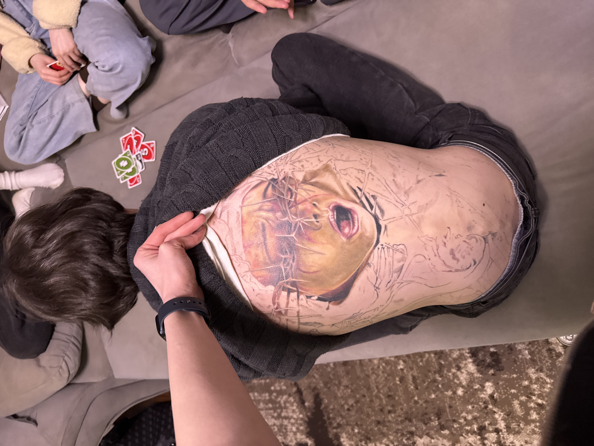 A person with a large, partially completed tattoo on their torso lies on a grey sofa. The tattoo depicts a stylized portrait with details including a face, possibly of Jesus Christ, with elements such as a crown of thorns. The person is wearing dark-colored denim pants, a grey sweater, and a white undershirt is visible. Another arm, wearing a black fitness tracker, extends towards the person's torso. To the left, partial views of other people are visible, including one person holding playing cards from the game Uno, with more cards scattered nearby. The carpet and surroundings suggest an indoor environment. The image was taken in Perm, Russia.