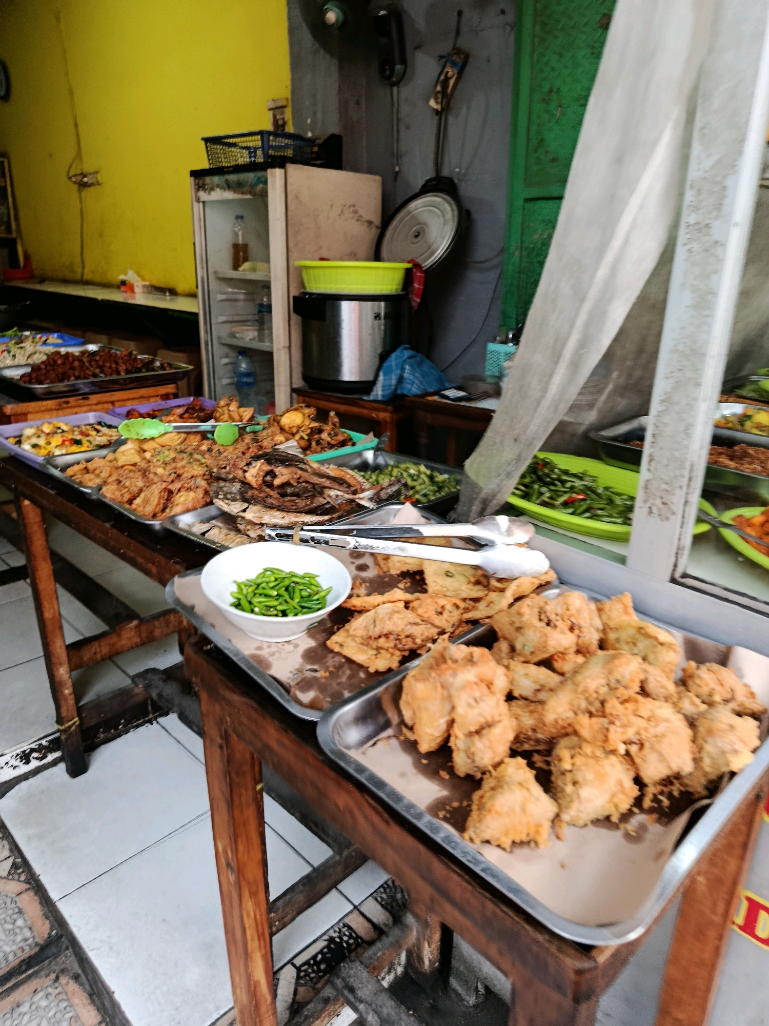 The image depicts an outdoor food stall with various Indonesian dishes displayed on wooden tables. The stall appears to be located in Bekasi, Indonesia, as indicated by signage that is partially visible with red and yellow text, common in Indonesian branding.
In the foreground, a metal tray is filled with golden-brown fried chicken pieces. To the left of the fried chicken, a white bowl contains small green chilies. Behind this tray, another metal tray holds an assortment of fried foods, including what appear to be pieces of tempeh and other fried items. Further back, more trays are visible, containing dishes such as fried fish, stir-fried vegetables (including green beans and possibly peppers), and other unidentifiable Indonesian cuisine. A green plastic container is also visible, as well as a metal pot.
The stall is set up against a building with a yellow wall on the left and a green door or wall section on the right. A white sheer curtain hangs from a frame, partially obscuring the view into the stall's interior. A refrigerator is visible in the background, along with a blue basket on top. The floor is tiled with a pattern of white and grey squares. The overall scene suggests a busy street food vendor environment. There are no people actively depicted in the image, but the setup implies recent or ongoing service. The lighting suggests it is daytime.
