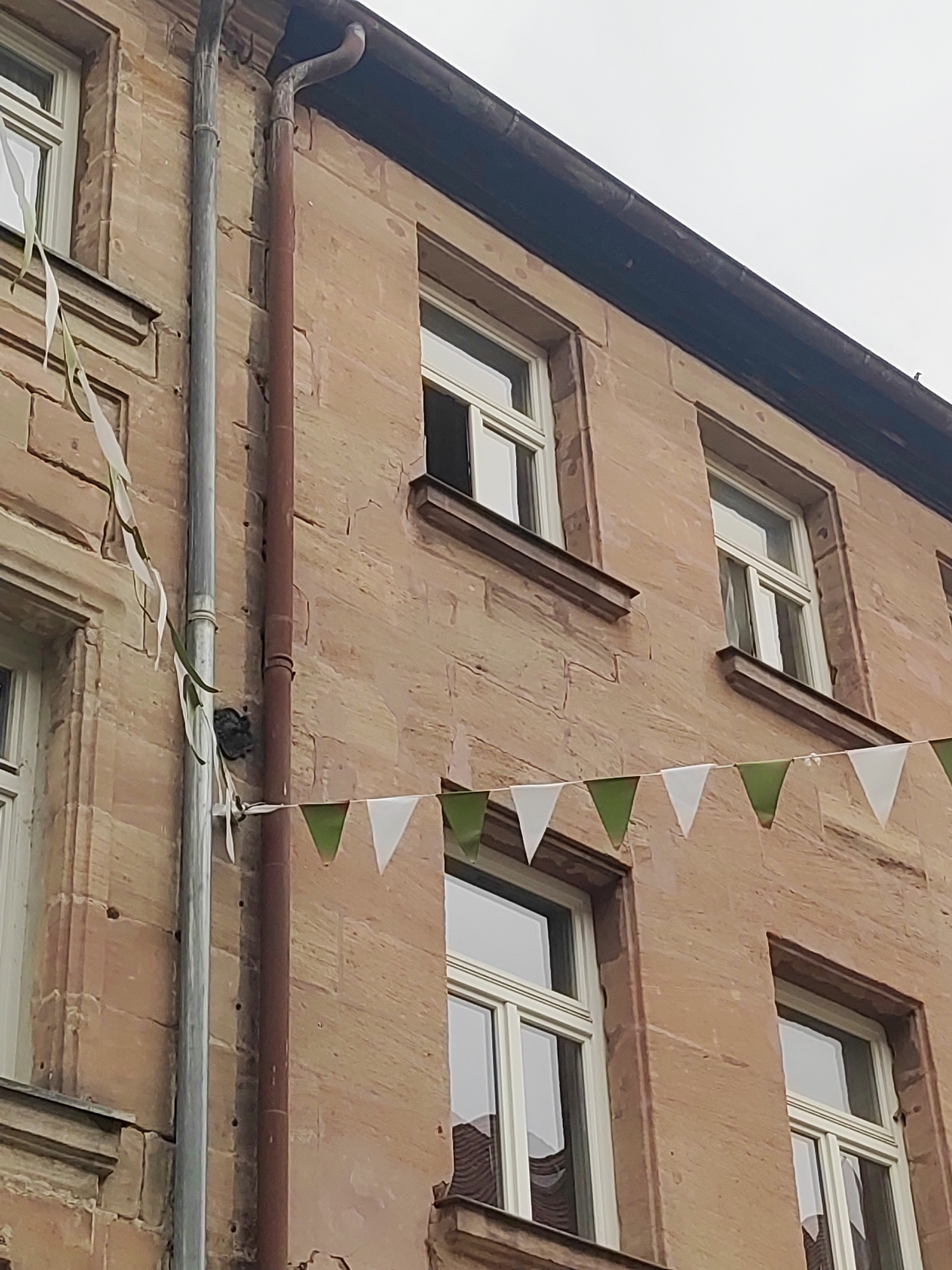 The image shows the facade of a building with sandstone walls. There are several windows with white frames visible. A rain gutter runs vertically down the left side of the building, and another, rust-colored, runs parallel to it. Bunting, made of alternating green and white triangular flags, is strung across the facade. There are also some white ribbons hanging down from the upper left. The sky is overcast. The image appears to be taken in Fürth, Germany, given the architectural style and the presence of a historic building.