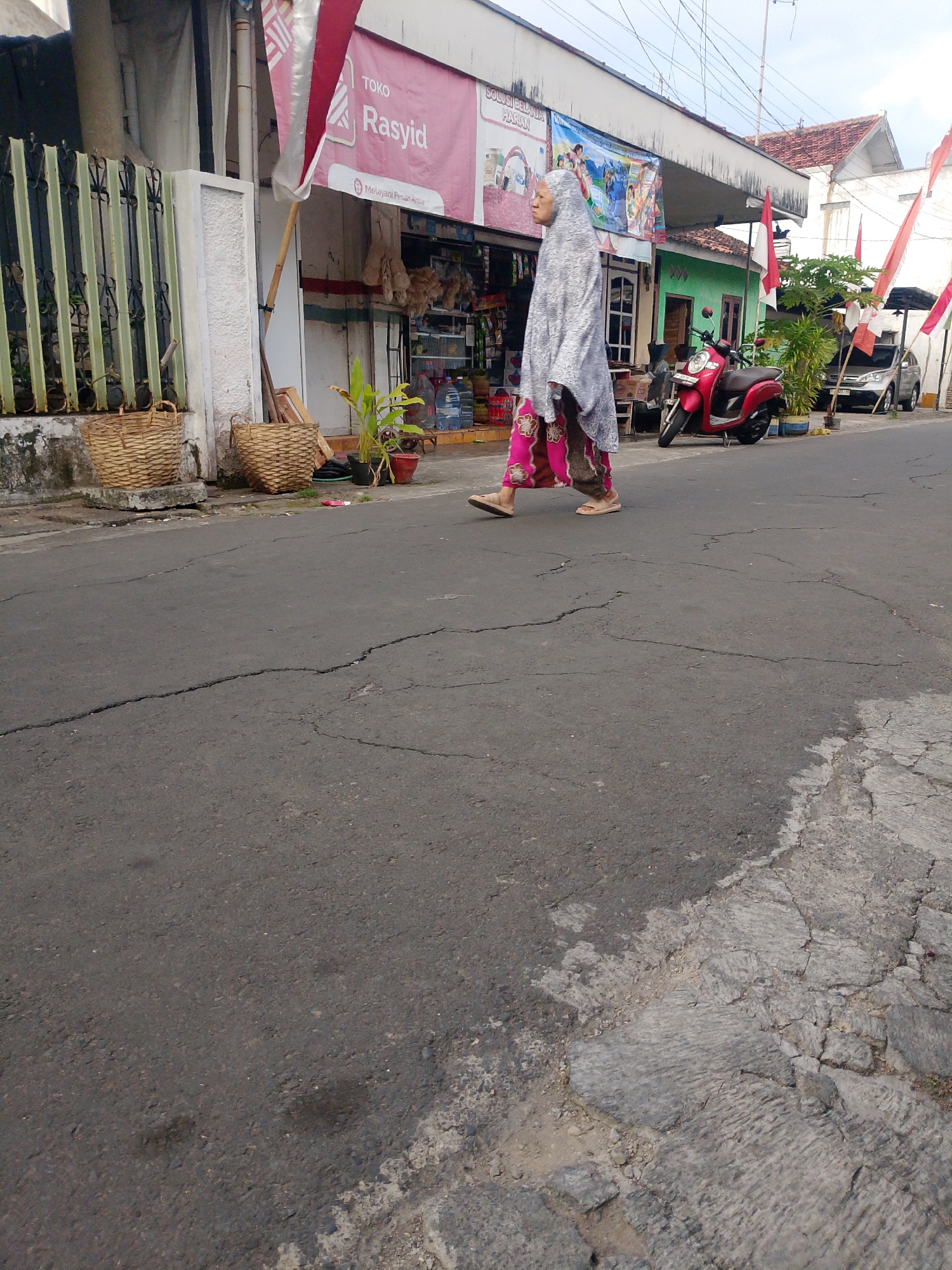 A full shot captures a street scene in Banyuwangi, Indonesia, with a focus on a woman walking. The woman, draped in a light-colored patterned hijab and a vibrant pink and patterned skirt, is depicted in mid-stride, her bare feet clad in simple tan sandals. She is walking towards the right side of the frame.
To her left, a row of shops lines the street. One shop, labeled "TOKO Rasyid," displays a red and white Indonesian flag. In front of the shops, two large woven baskets sit on the sidewalk. A potted plant adds a touch of greenery to the scene.
Further down the street, a bright red scooter is parked. Beyond the scooter, a silver car is visible. The street itself is paved with asphalt, showing signs of wear and tear with visible cracks. The environment appears to be an urban setting with buildings and a cloudy sky overhead. The overall impression is that of a typical day in a bustling Indonesian town.