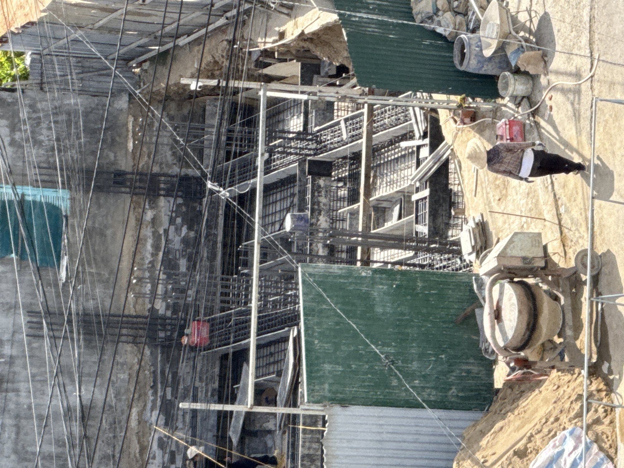 The image depicts an outdoor construction site in Bac Nha Trang Ward, Vietnam. In the foreground, a person wearing a conical hat, plaid shirt, dark pants, and white gloves walks away from the camera. Beside them is a cement mixer and a pile of sand. Further back, a green corrugated metal fence stands prominently. The background shows a multi-story structure under construction, with exposed rebar and concrete forming the framework of the building. Electrical wires crisscross the scene, strung across the gray concrete wall. A bright red object is visible on an upper level of the construction. The overall impression is of an active, ongoing construction project under daylight.