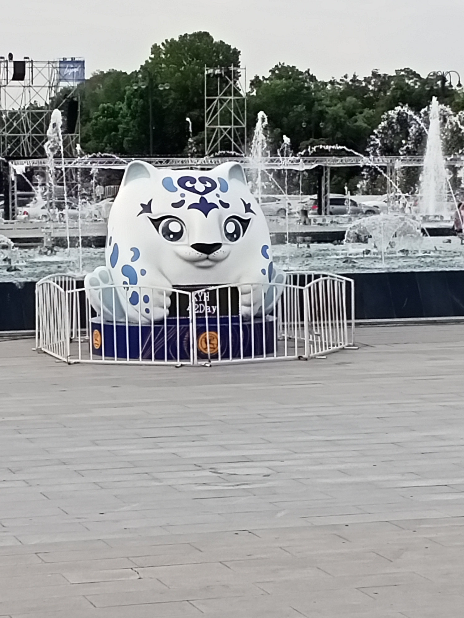 A large, round, white statue of a snow leopard with blue spots and markings sits behind a white metal fence. The statue is in front of a fountain and trees. There are also some large metal structures in the background, which may be part of a stage or event setup. A sign on the statue base reads "KYH 42 Day". The area appears to be a public outdoor space, possibly a park or plaza in Bishkek City, Kyrgyzstan. The ground is paved with large gray tiles. The sky is overcast.