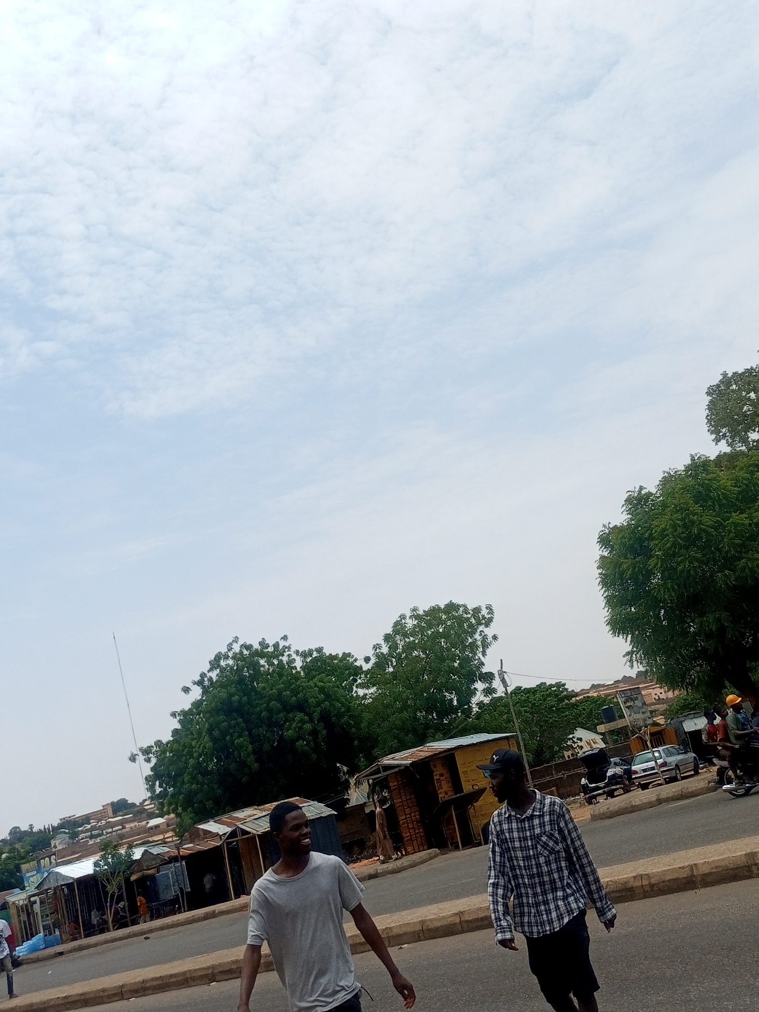 Two young men are walking on a street in Tempure, Nigeria. One man is wearing a gray t-shirt and jeans. The other man is wearing a black and white plaid shirt, a black cap, and shorts. They appear to be in conversation. Behind them, there are street-side shops. Cars are parked along the street. Trees dot the landscape. The sky is overcast.