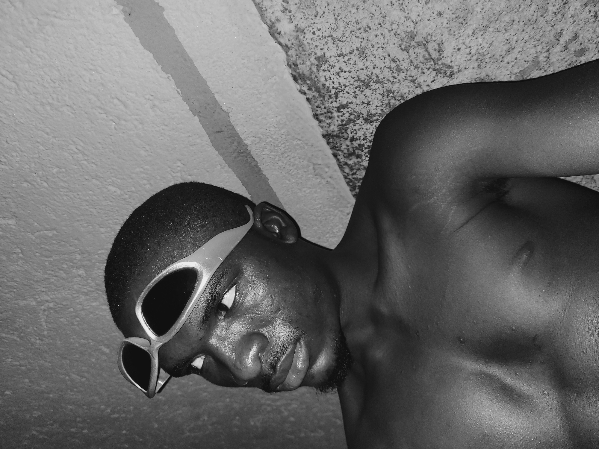 The black and white, close-up, waist-up shot shows a young Black male looking up and to his left, away from the camera. He has short, dark hair and a goatee. He is wearing clear-lensed swimming goggles pushed up onto his forehead, with the straps going around his head. His skin appears moist, possibly from sweat or water. He is shirtless, revealing his chest and shoulder. The background consists of a textured, light-colored wall with a diagonal stripe of a slightly darker shade. To the right of the image, a rough, textured surface, possibly a concrete wall or ground, is visible. The lighting casts a shadow from a horizontal element onto the wall behind the subject. The overall setting suggests an indoor or sheltered outdoor location in Bukuru, Nigeria, possibly a bathroom or poolside area, given the swimming goggles. There are no other individuals present in the frame.