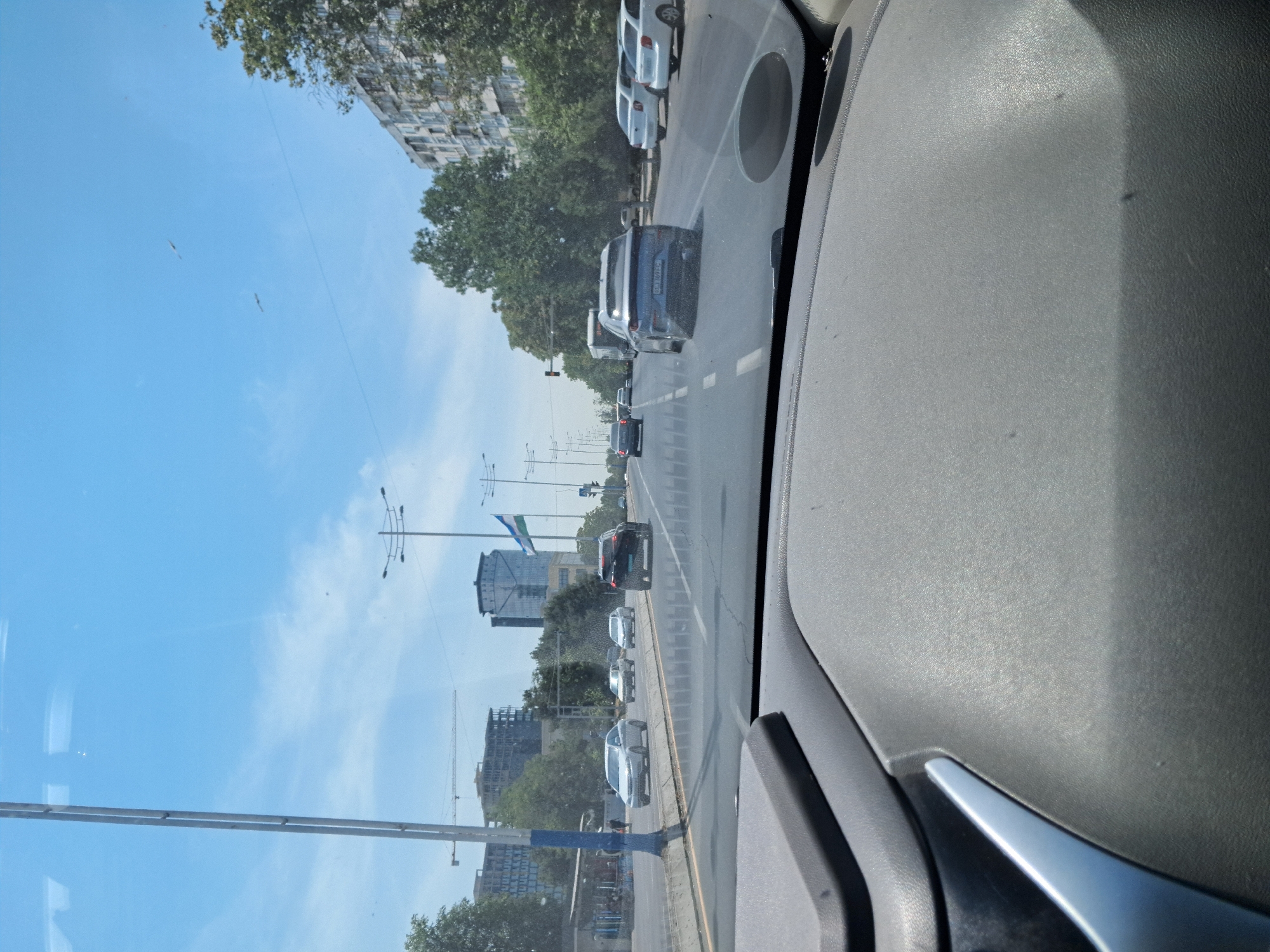 The image is taken from inside a vehicle, looking out through the windshield at a city street in Tashkent, Uzbekistan. The sky is clear blue with a few wispy clouds. Several cars are driving on the multi-lane road, including a black SUV, a blue sedan, and several white sedans. On the side of the road, there are trees, buildings, and streetlights. A construction crane is visible in the distance, along with several multi-story buildings. A flag is flying from a pole in the middle ground. The foreground shows the dashboard of the car.