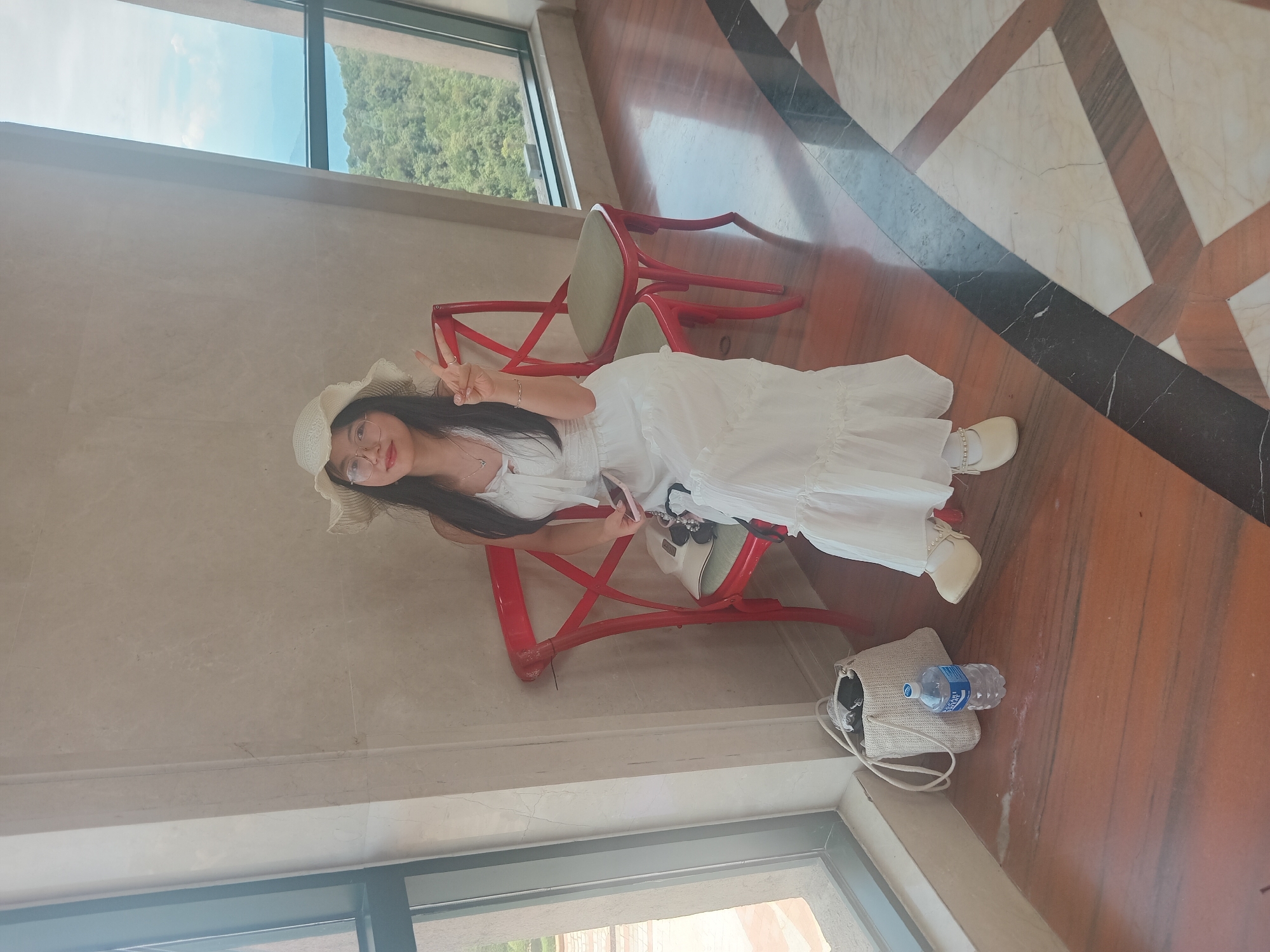 A young Asian woman wearing a white sundress and a straw hat is seated on a red chair in front of a large window. She is holding a smartphone in her right hand and making a peace sign with her left hand. A white purse and a bottle of water are on the floor next to her. The background shows a view of lush green mountains and a cloudy sky. The floor is made of polished wood with a marble inlay pattern. This scene is likely located at a restaurant or cafe with a scenic view, possibly in the Ba Na Hills area outside of Đà Nẵng, Vietnam.