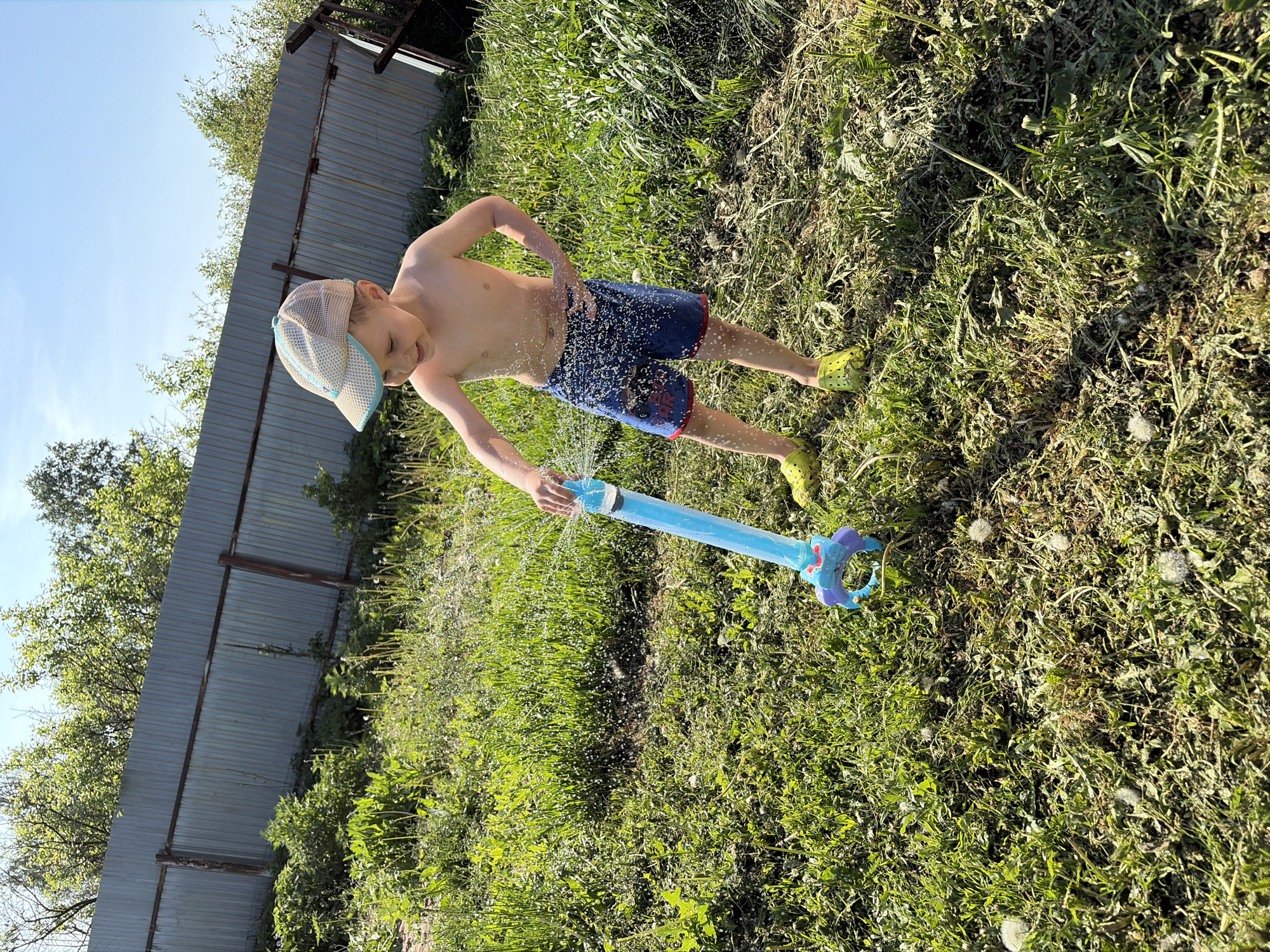 A boy in a light-colored cap, blue swim trunks with a red stripe, and chartreuse footwear stands in a grassy area, operating a blue water squirter. He is barefoot. The grass appears untended, with cut sections intermingled with standing growth. A metal fence borders the grassy area. Trees are visible beyond the fence. The scene is well-lit, suggesting daylight conditions. The location appears to be in a rural area in Фоки, Russia.