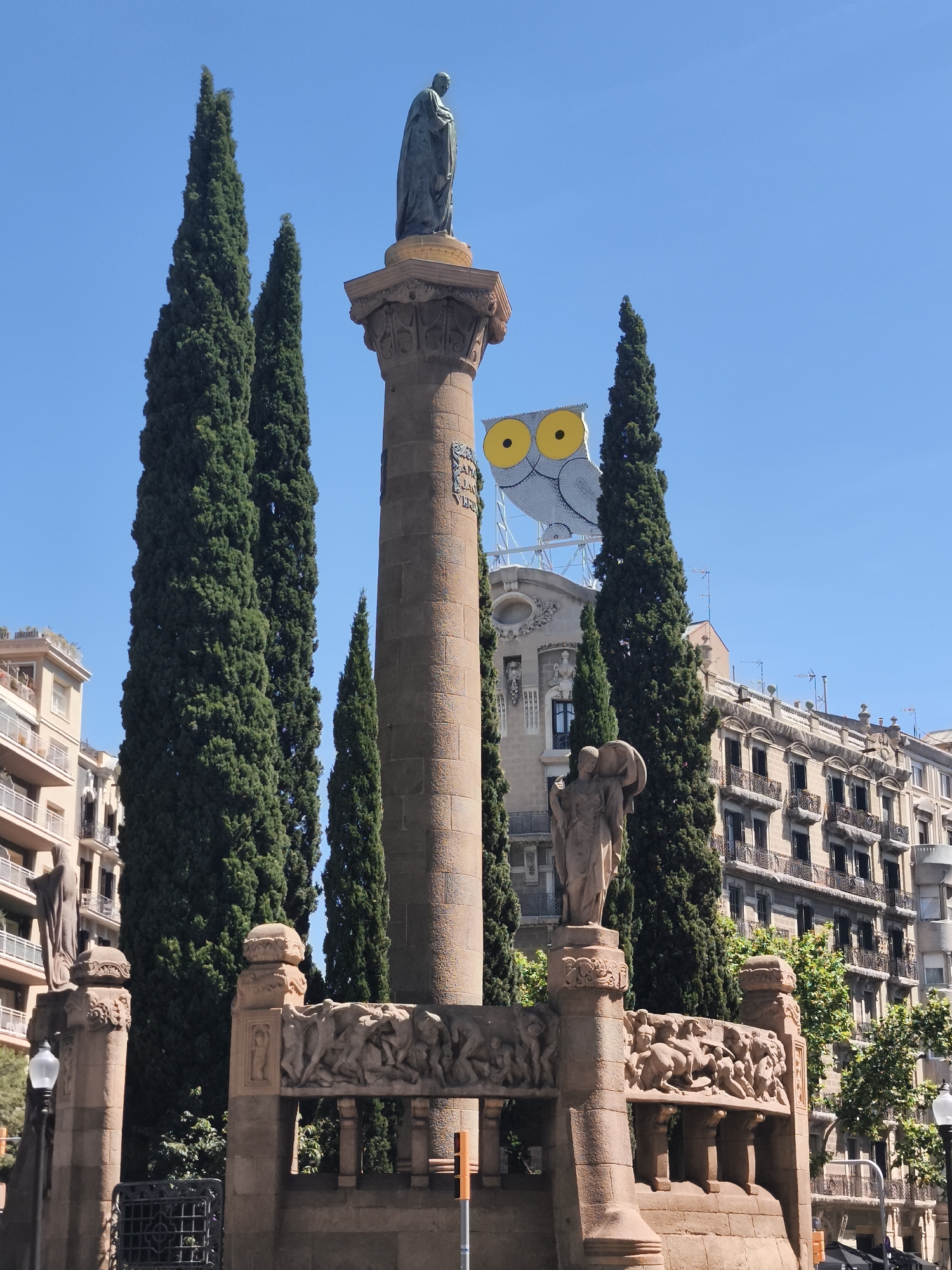 The image depicts the Column of Joan Prim, a monument located in the Plaça de la Ciutadella in Barcelona, Spain. The monument consists of a tall, cylindrical stone column topped with a statue of Joan Prim, a 19th-century Spanish general and statesman. The base of the column is decorated with sculpted figures. The monument is surrounded by tall, slender cypress trees. In the background are multi-story buildings, one of which has a large advertisement featuring a cartoon owl with prominent yellow eyes. The sky is blue.