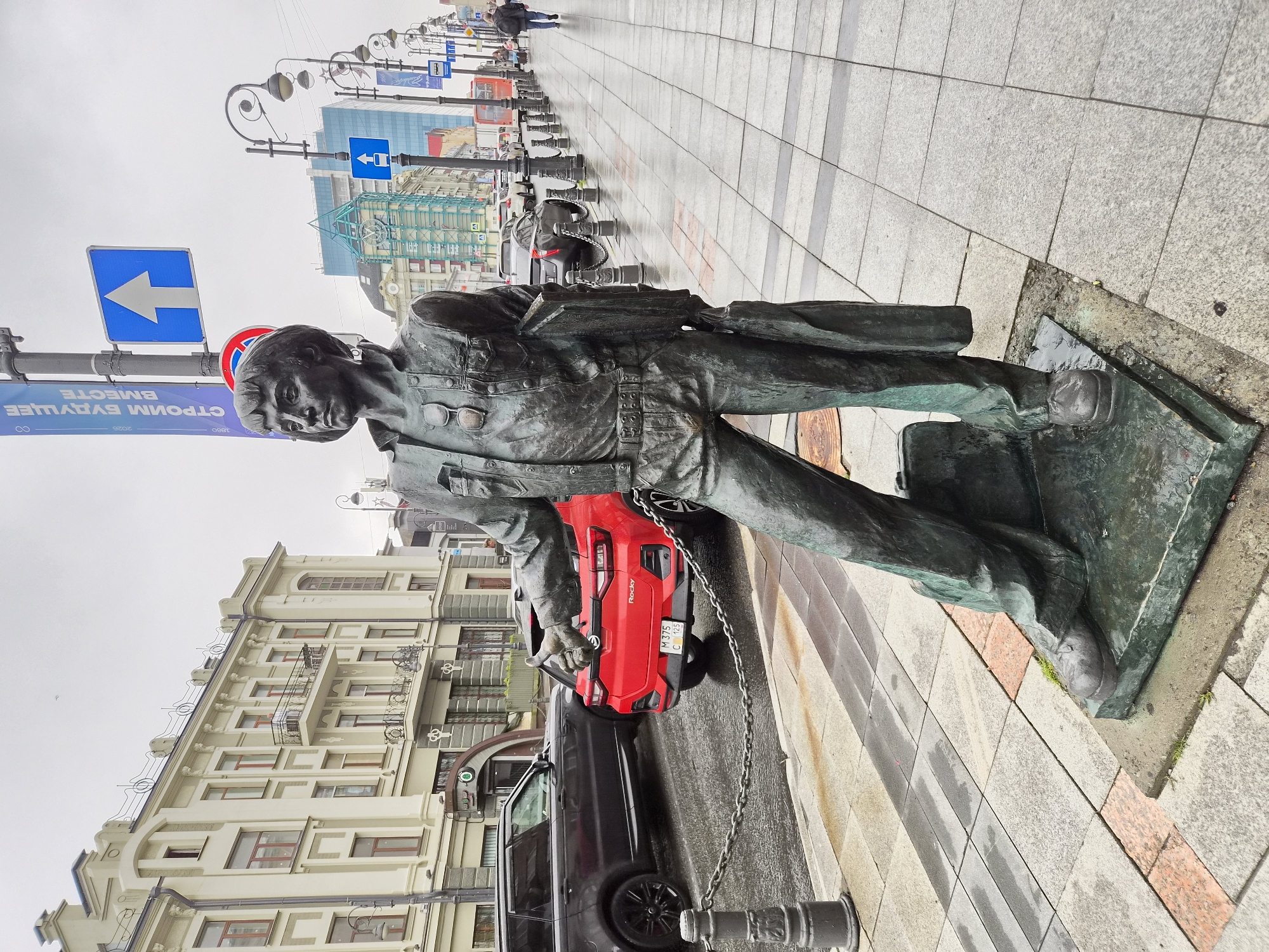 The photograph captures a bronze statue of a person standing on a paved sidewalk in Vladivostok, Russia. The statue, rendered in a realistic style, depicts a figure wearing a jacket, shirt, belt, and trousers, with a briefcase in one hand and a book or folder in the other. Sunglasses are perched on the statue's chest. The figure's right thumb is extended upwards in a "thumbs-up" gesture.
The statue is positioned on a raised, grey-green metal base, which itself rests on a stone plinth. The surrounding pavement is laid with rectangular and square stone tiles, some of which are wet and reflective, indicating recent rain. A chain-link fence with bollards runs alongside the sidewalk, separating it from the street.
In the background, a dark grey SUV is parked on the left, and a red hatchback car is positioned behind it, with its rear facing the camera. The red car has a license plate that reads "M 375 C 125".
Further down the street, a row of lampposts with decorative scrolling designs lines the sidewalk. Several buildings are visible, including a light-colored, multi-story structure with ornate architectural details and red-trimmed windows on the left, and a modern building with a blue-tinted glass facade further down on the right. Traffic signs, including a blue sign with a white arrow pointing upwards, are also present.
A few other individuals are visible in the distance on the sidewalk, walking away from the camera. The sky is overcast and grey. The overall scene suggests an urban environment on a damp day.