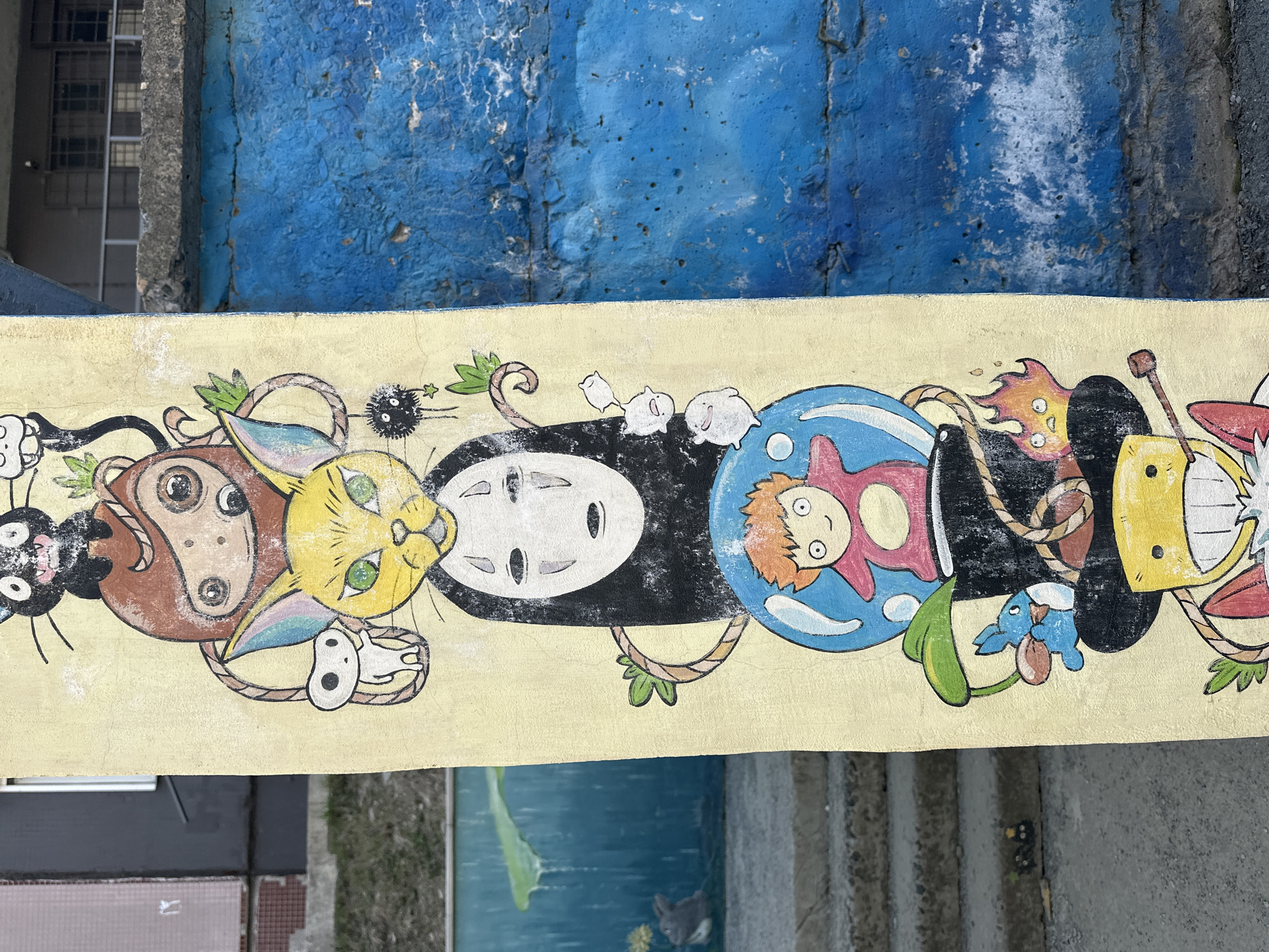 This media file displays a vibrant mural featuring popular characters from Studio Ghibli films, painted on a light yellow-beige wall. The mural is oriented horizontally, depicting a sequence of characters seemingly connected by green leafy vines and brown rope-like elements.
From left to right, the mural shows Jiji, the black cat from *Kiki's Delivery Service*, followed by a brown Totoro with large eyes, and then the yellow Catbus, both from *My Neighbor Totoro*. Next is No-Face from *Spirited Away*, rendered with its iconic white mask and black body, surrounded by small black soot sprites (susuwatari). Further right, Ponyo, the red-haired girl, is depicted inside a blue bubble or container. Interspersed are small white blob-like creatures, possibly Kodama or additional soot sprites, and a small blue creature. The final character on the right is a stylized yellow figure with large teeth and reddish details.
Above the mural, a rough, heavily textured wall is visible, painted in varying shades of blue, showing signs of wear and peeling paint. Below the mural, the image hints at a different surface, possibly ground or another wall, with suggestions of water in some areas. The lighting suggests an outdoor setting, though the time of day is not discernible. No text is visible in the image. The overall impression is one of whimsical street art.