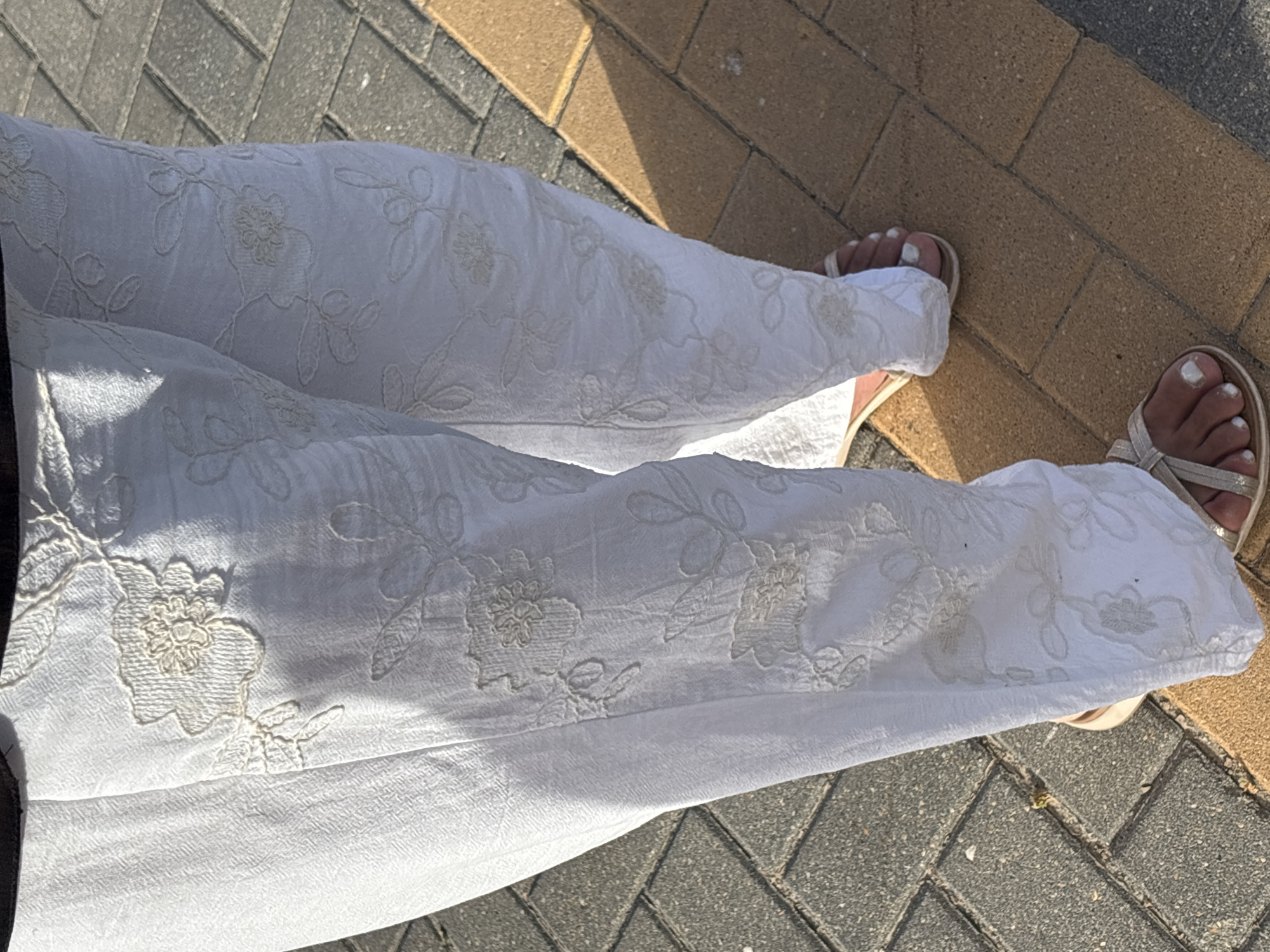 The image shows a close-up view of a person's legs wearing white embroidered pants and sandals, standing on a brick-paved sidewalk in Urbanización Cabo Roig, Spain. The pants have intricate floral embroidery in a lighter shade of white. The sidewalk consists of grey and tan rectangular bricks. The lighting suggests it is daytime, with a shadow cast on the pavement. The scene is depicted from a low angle, focusing on the lower half of the body and the ground.