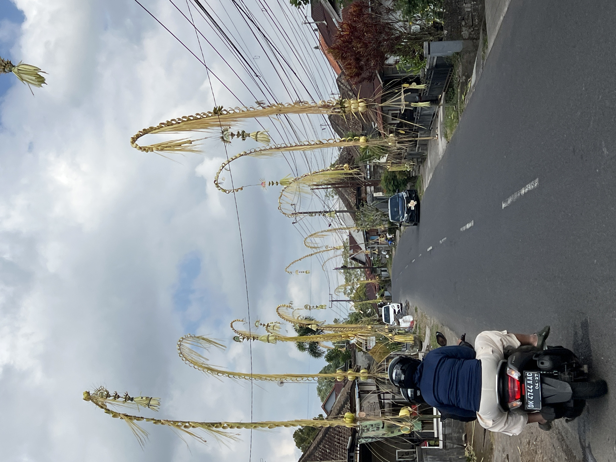 In the city of Pupuan, Indonesia, a street is adorned with tall, decorative palm leaf structures, known as "penjor," which are characteristic of Balinese culture and often displayed during holidays and ceremonies. These "penjor" are positioned along the sides of the road, with some arching over the street. The street itself is paved and appears to be a moderately trafficked road, with at least one car and a scooter visible. The houses lining the street have traditional tiled roofs and are set back from the road. Overhead, a network of electrical wires crisscrosses the sky, and the weather appears to be overcast with scattered clouds. In the foreground, a person is riding a scooter, facing away from the camera, wearing a blue jacket and light-colored pants. The license plate of the scooter is visible and reads "DK 4744 AAC". The overall scene depicts a streetscape with cultural decorations, residential buildings, and everyday transportation in a tropical setting.