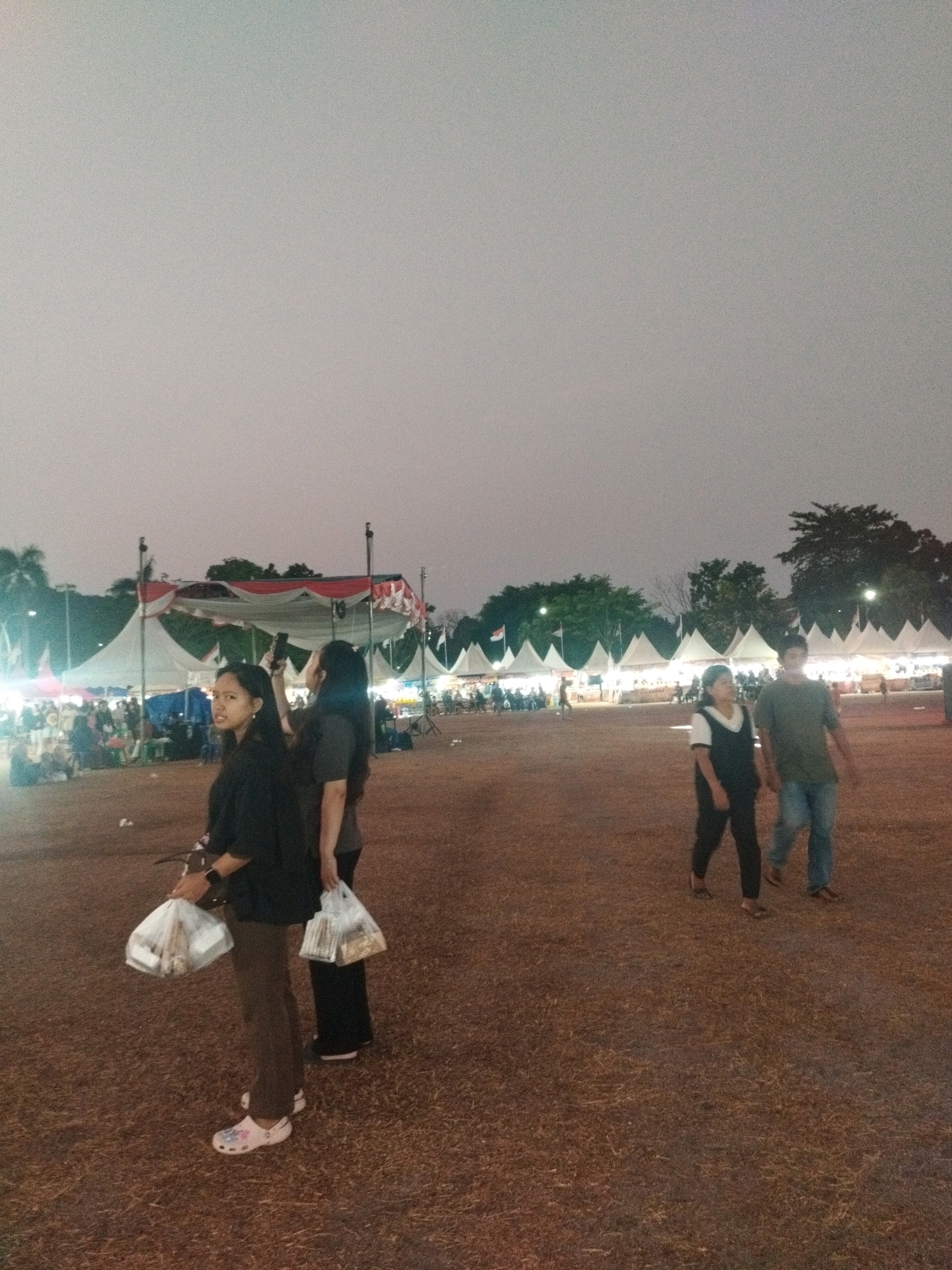 The image depicts an outdoor event, possibly a fair or market, in the city of Parit Padang, Indonesia. The sky is overcast and the time appears to be late afternoon or early evening. In the foreground, two young women are walking away from the camera. The woman on the left is wearing a black long-sleeved shirt, brown pants, and white clogs adorned with floral patterns. She is carrying two white plastic bags in her left hand. The woman next to her, on the right, is wearing a dark grey t-shirt and dark pants. She is looking back towards the left, seemingly at the woman beside her or something behind them.
Further in the background, there is a row of tents, likely stalls for vendors, with lights illuminating them. Several people can be seen gathered around these tents. Another couple is walking from right to left in the mid-ground. The man is wearing a green t-shirt and blue jeans, while the woman beside him wears a black vest over a white shirt and black pants. The ground is covered with dry grass and debris. Trees are visible in the distance beyond the tents. The overall impression is of a bustling public gathering.