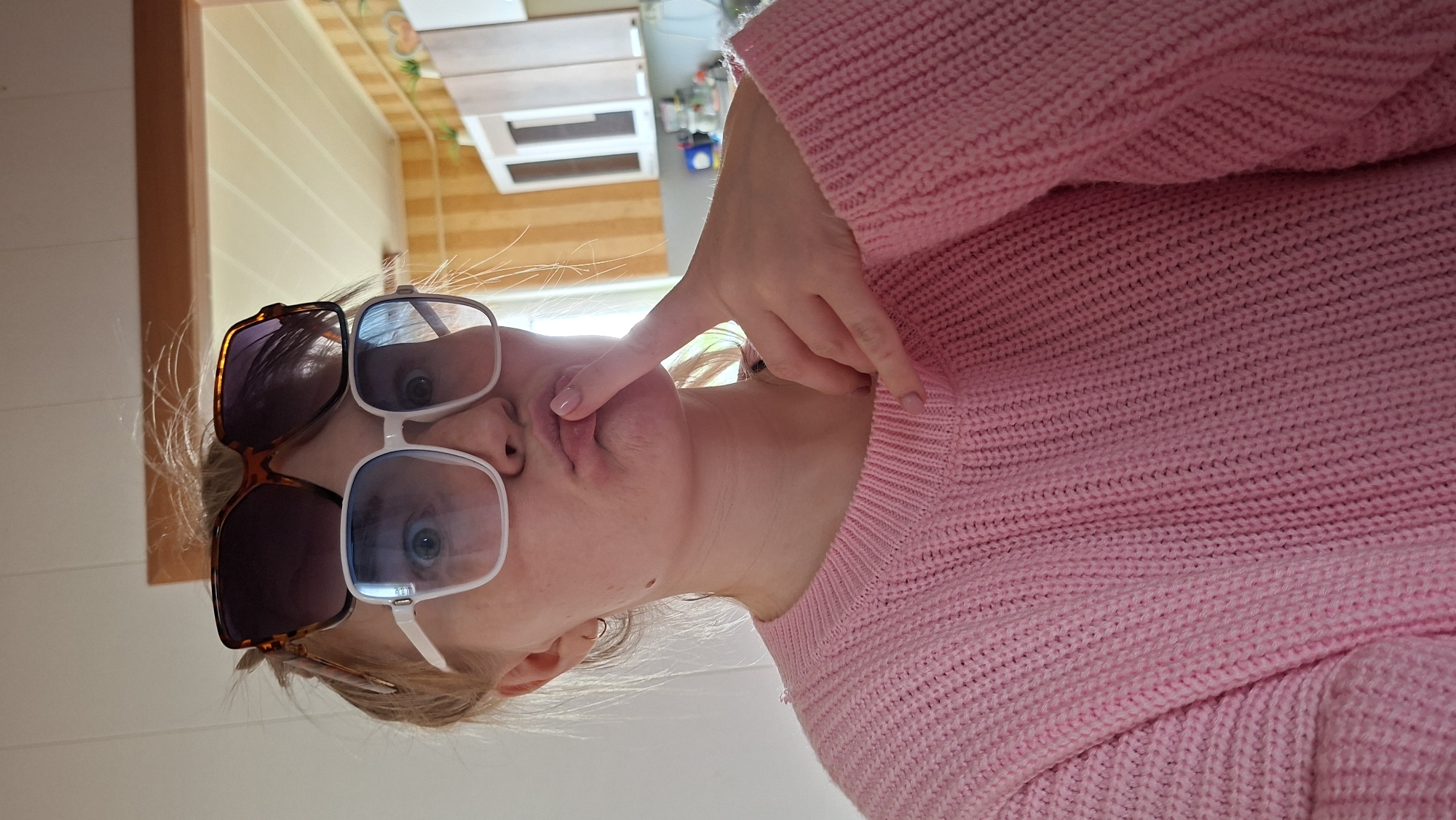 The image depicts a woman wearing two pairs of sunglasses, one tortoiseshell-framed pair stacked atop a white-framed pair. She is wearing a pink knitted sweater. Her right hand is raised, with a finger touching her pursed lips. The background includes a light-colored wall and glimpses of a wooden structure, possibly furniture or part of the interior architecture. The image is taken from a slightly low angle, with the subject appearing to look down at the camera. The location is not discernible from the image.
