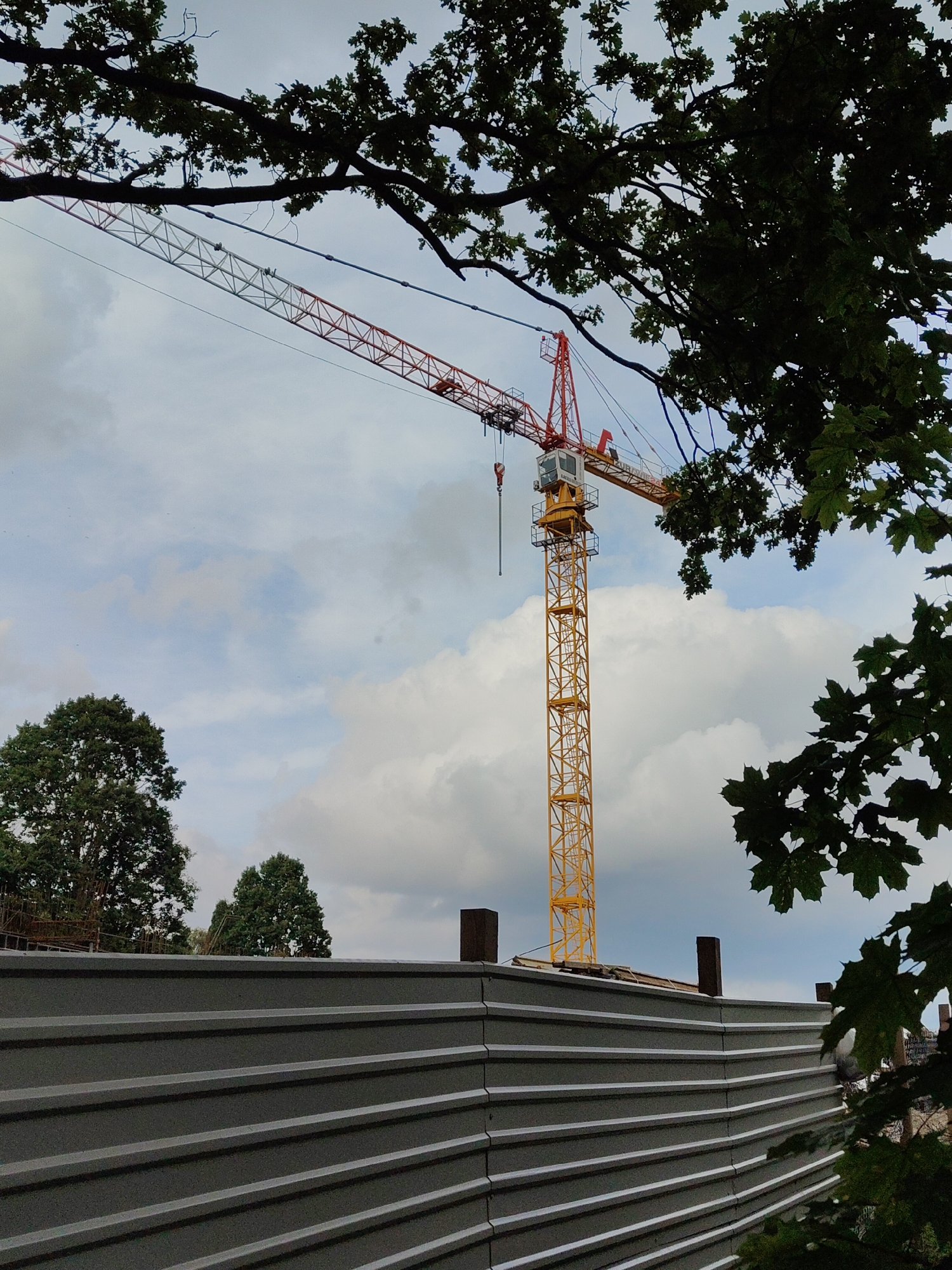 An outdoor, slightly low-angle medium shot shows a yellow construction crane against a cloudy sky, partially framed by the dark green leaves and branches of trees. The crane's tower is a lattice structure painted yellow, and its boom extends diagonally across the frame, with sections painted white and red. A control cabin is visible near the top of the tower. In the foreground, a corrugated metal fence runs horizontally across the bottom third of the image. Behind the fence and to the left of the crane, the tops of two mature green trees are visible. The environment appears to be a construction site in the city of Rumia, Poland, possibly during daylight hours given the overall brightness of the scene.