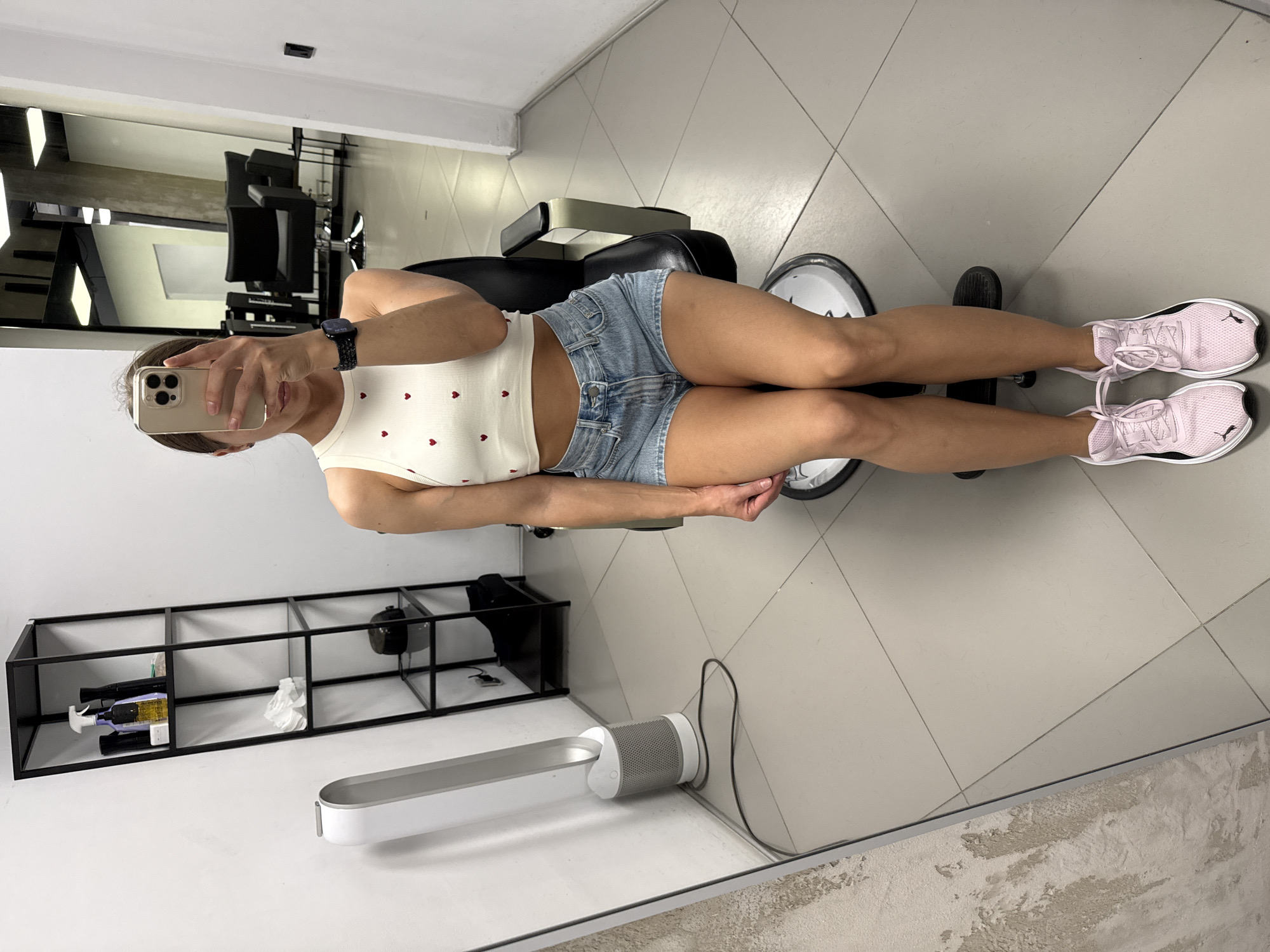 A woman is taking a mirror selfie in a salon. She is wearing a white crop top with red hearts, denim shorts, and pink sneakers. She is sitting in a black salon chair. To her left is a white Dyson fan and a black shelf unit containing various bottles and objects. In the background, there are more salon chairs and mirrors. The flooring is tiled. The scene is indoors, likely in a salon or barbershop, possibly located in Saint Petersburg, Russia, based on the context provided.
