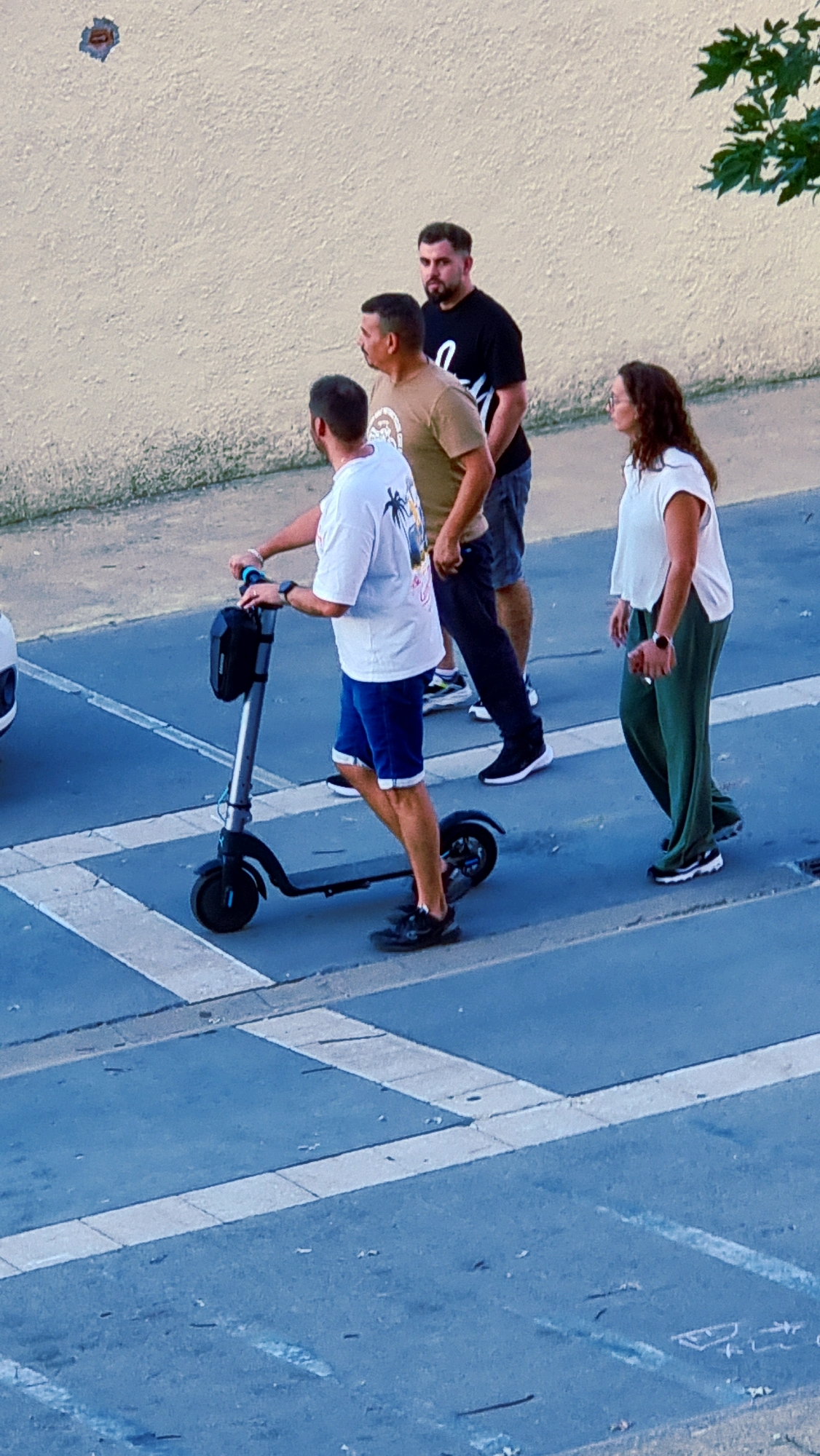 In Sant Joan Les Fonts, Spain, four individuals are present on a paved surface that appears to be a street or parking lot with painted white lines. A man in the foreground is riding a grey electric scooter, wearing a white t-shirt with a palm tree graphic and blue shorts. He has dark hair and appears to be looking forward. Behind him, two men are walking. The man in the middle is wearing a tan t-shirt and dark shorts. The man in the back is wearing a black t-shirt. To the right, a woman is walking, wearing a white t-shirt and green pants. She is looking to her right. The background consists of a textured, light-colored wall and some green foliage on the upper right.