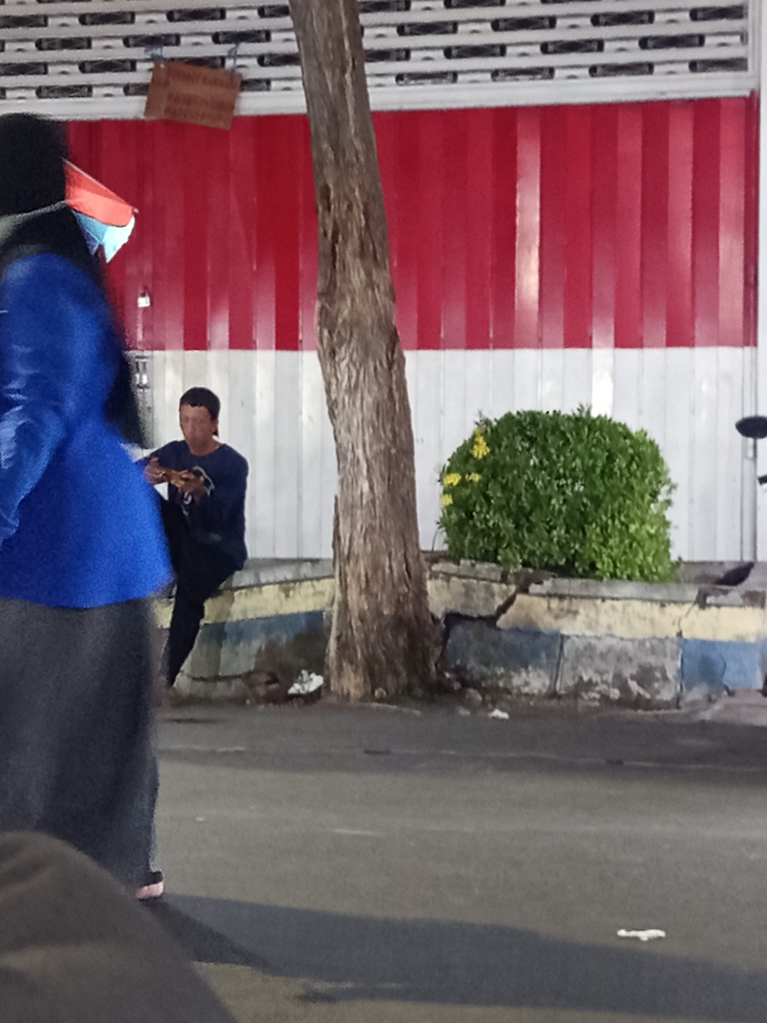 The image captures a street scene in Situbondo, Indonesia. In the foreground, a person wearing a blue jacket and a grey skirt is partially visible, with a red and blue patterned mask partially obscuring their face. To the left, a small wooden sign is attached to a rolling shutter door, which is painted with horizontal red and white stripes. In the middle ground, a person in a dark blue long-sleeved shirt and dark pants is seated on a concrete bench, appearing to eat or hold something small in their hands. Behind them, a dense green bush with yellow flowers is planted in a raised planter box. A large tree trunk is positioned between the seated person and the viewer. The background consists of a rolling shutter door and a white tiled wall. The ground appears to be asphalt or pavement. The lighting suggests it is daytime.