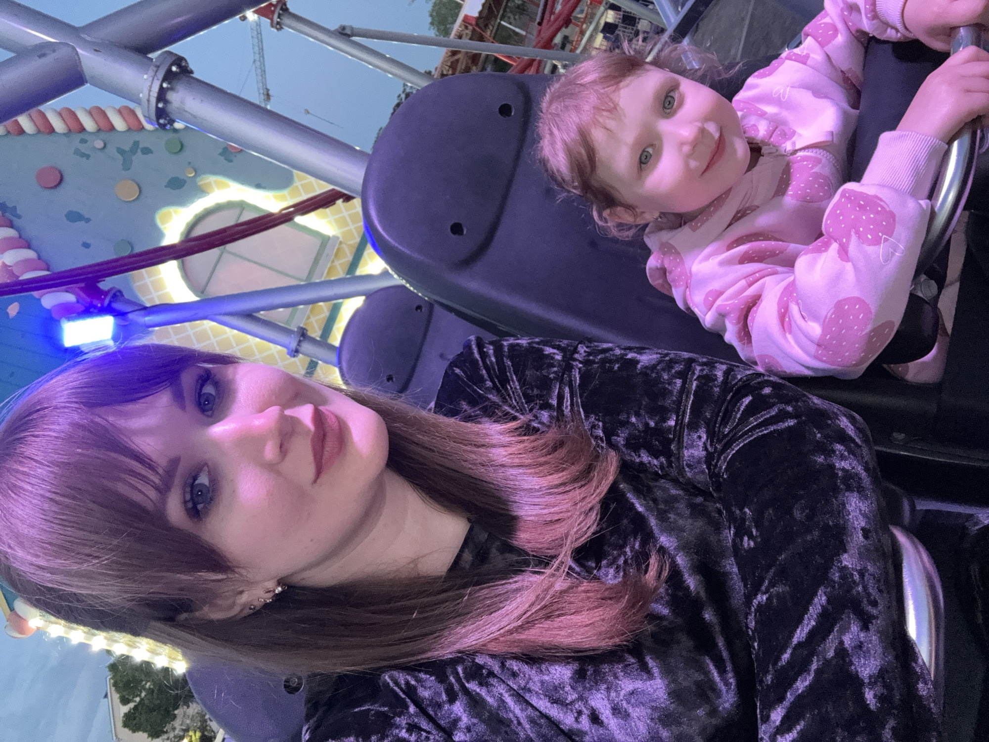 The photograph, taken at an oblique angle, depicts a woman and a young child on an amusement park ride, likely located in Yevpatoriya, Ukraine. The woman, wearing a black velvet-like top and sporting auburn hair with bangs, is positioned in the foreground. Her features include light eyes and lipstick. The child, wearing a pink strawberry-patterned sweatshirt, is seated in the ride beside her, grasping the safety bar. The ride is constructed with metal pipes and a seat back. In the background, a structure resembling a colorful, cartoon-themed building can be seen, along with hints of amusement park lighting and other rides. The overall illumination suggests the photo was taken during twilight or at night.
