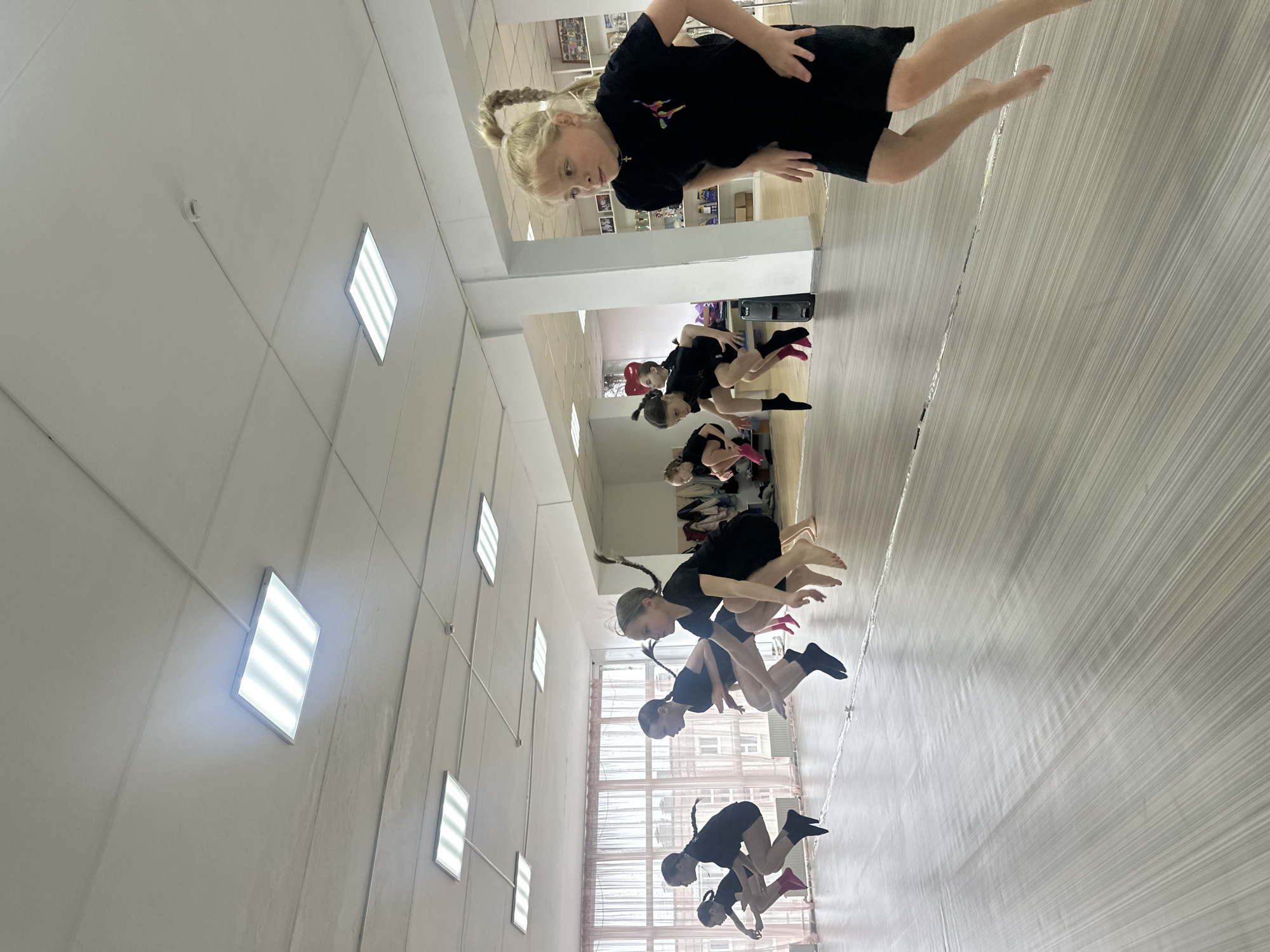 The image shows a group of girls in a dance studio. The girls are wearing black dresses and some have socks or shoes on. They appear to be practicing jumps or leaps. The studio has a row of rectangular lights on the ceiling and is located in Votkinsk, Russia.