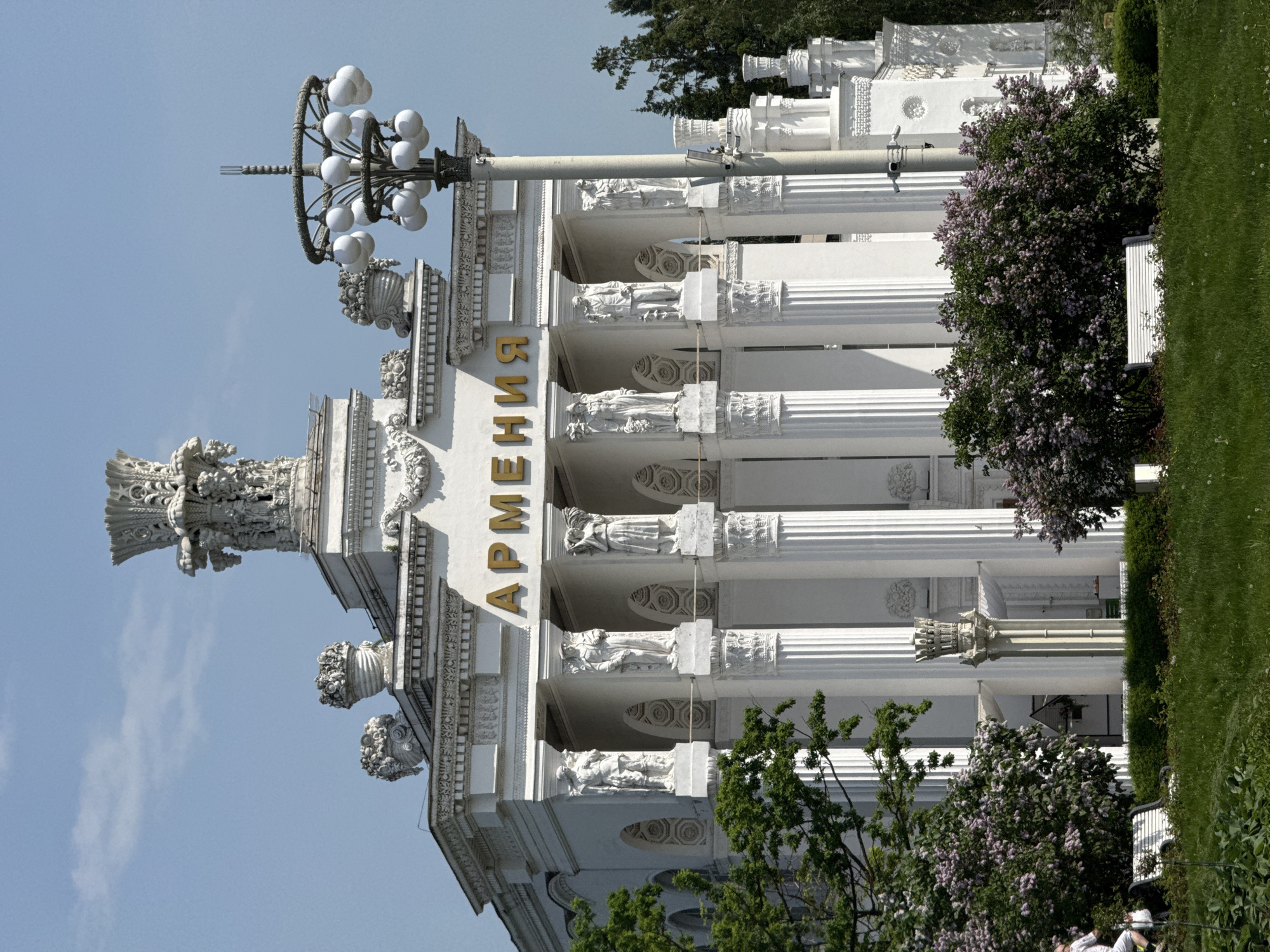 The image shows the Armenia Pavilion at the Exhibition of Achievements of National Economy (VDNKh) in Moscow, Russia. The white facade features classical architectural elements, including columns, statues, and ornate details. The word "Армения" (Armenia) is displayed vertically in gold lettering. A decorative light fixture stands to the left. Trees with green and purple flowers frame the building. A patch of green lawn is visible to the right. The sky is a clear, light blue.