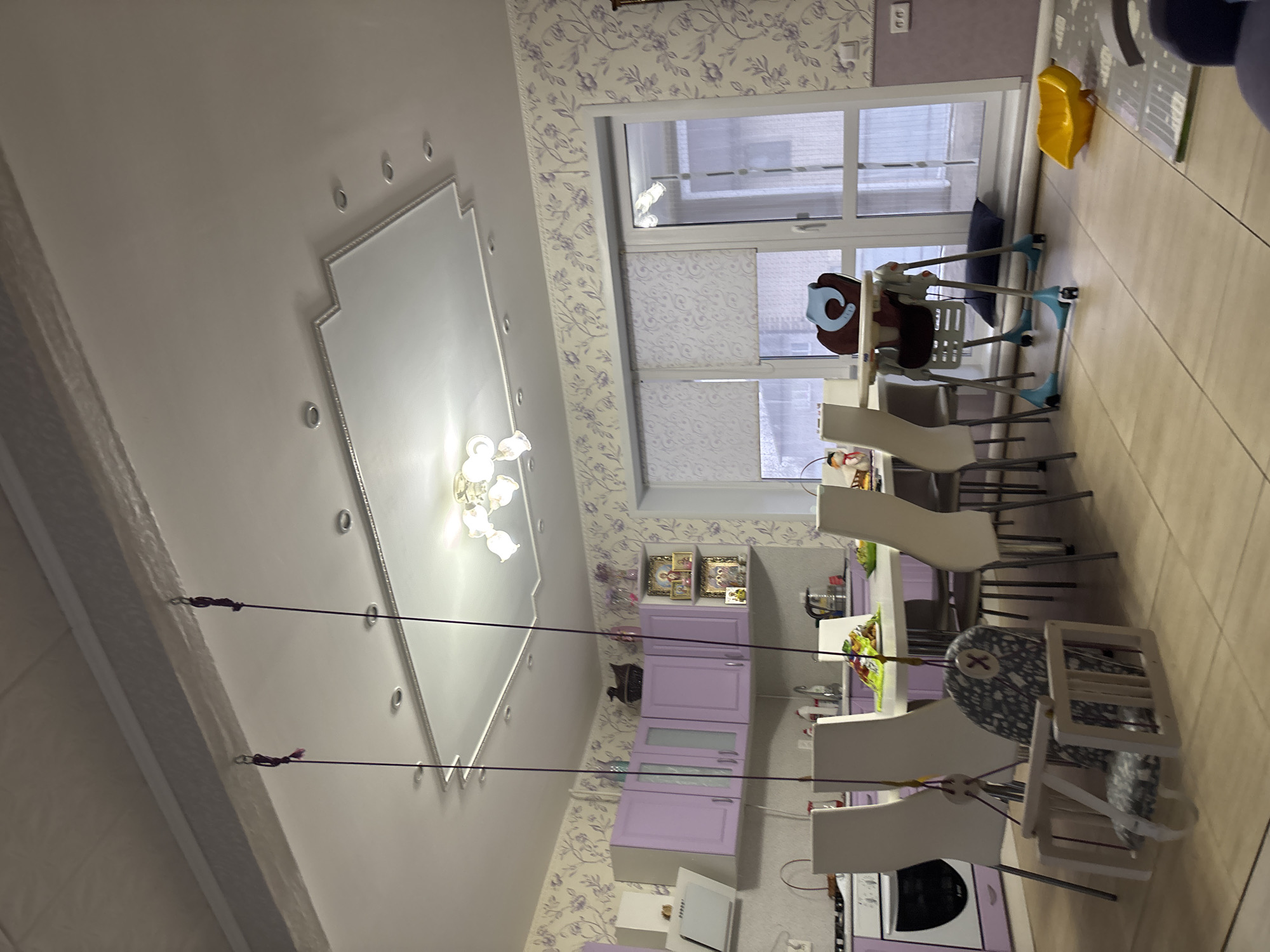 A tilted medium-shot view in a kitchen. The wall and part of the ceiling are visible. The wall is wallpapered with a purple floral design. The ceiling is white and has a decorative recessed panel with a chandelier. There is a window in the upper right of the image with a light-colored shade. The window is adjacent to a tiled floor. In the right side of the image, there are chairs and a high chair. There is also a kitchen with light purple cabinets. The kitchen has a sink, countertops, and appliances. There is also a swing hanging from the ceiling. It's unknown if this photo was taken in Ферма, Russia.