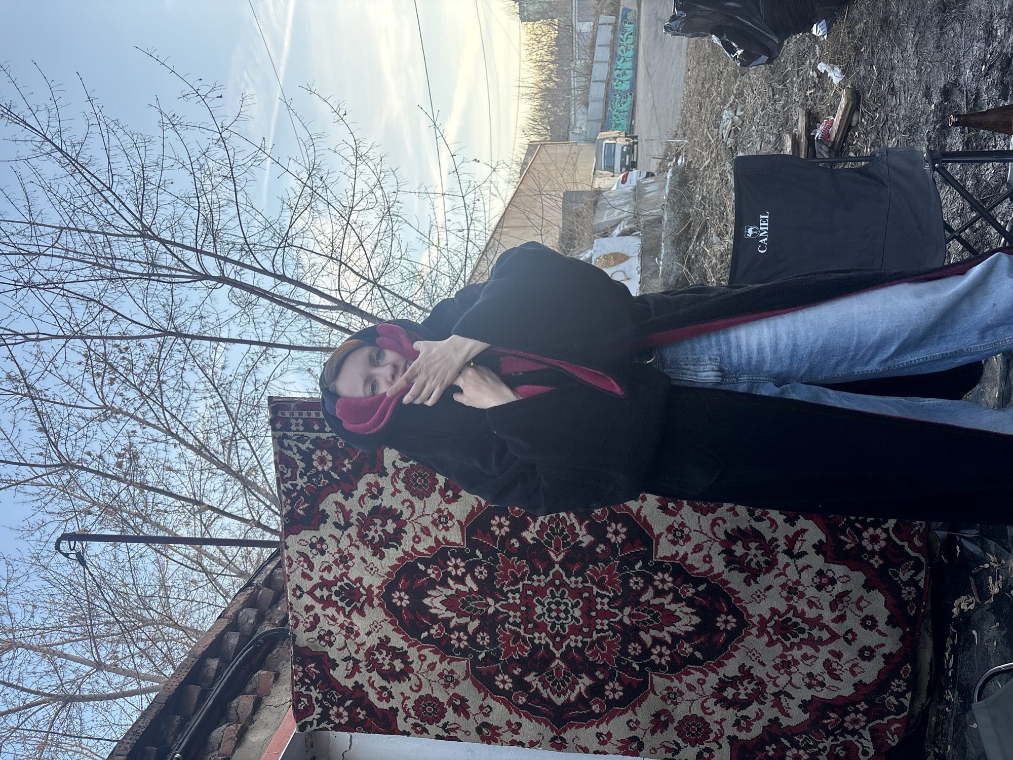 A person in a black coat and blue jeans stands outdoors next to a carpet with a red and beige floral pattern. The person's face is partially obscured by their hands and a pink hood. Behind them is a black folding chair with the word "CAMEL" printed on it. The ground is littered with debris, including trash bags and discarded items. In the background, there is a building with graffiti and a light-colored sky with thin clouds. The location appears to be in an urban area. Based on the visual cues of architecture, urban waste, and general environment, this may be Krasnoyarsk, Russia. Bare tree branches are visible in the upper left corner.
