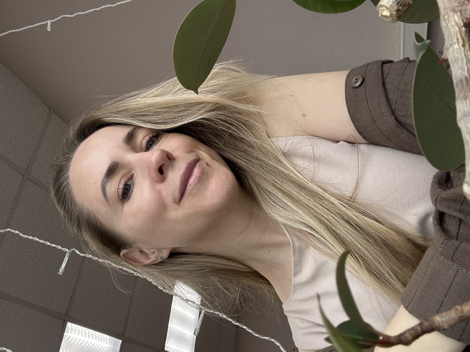 A woman with long blonde hair is positioned at the bottom of the frame, looking upwards. She wears a light-colored top and a brown sleeve with button detail. A plant with large green leaves occupies the right side of the frame, obscuring the background. A string of lights is draped near the top left. The ceiling is visible, with a grid pattern and fluorescent lighting, indicative of an office environment. The location is Chaikovsky, Russia.