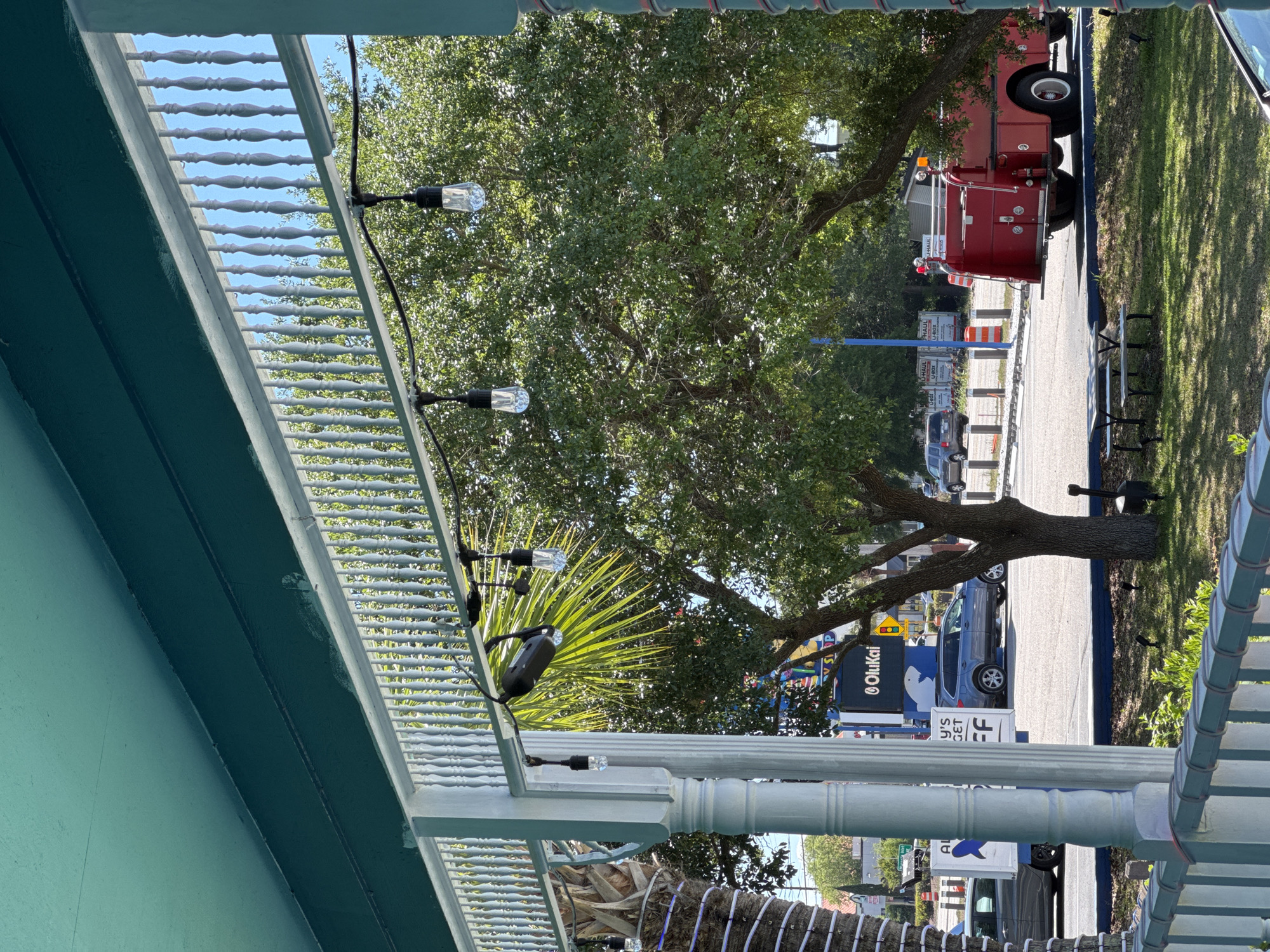 The image shows a partial view from under a porch or balcony, featuring a green-painted structure with white railings adorned with string lights. The scene outside captures a street in Garden City Beach, United States of America. A large tree dominates the view, partially obscuring a fire truck parked along the road. Various parked vehicles and street signs, including one for "OluKai", are visible along the roadside. Additional features include landscaping, walkways, and glimpses of nearby buildings. The lighting indicates a sunny day.