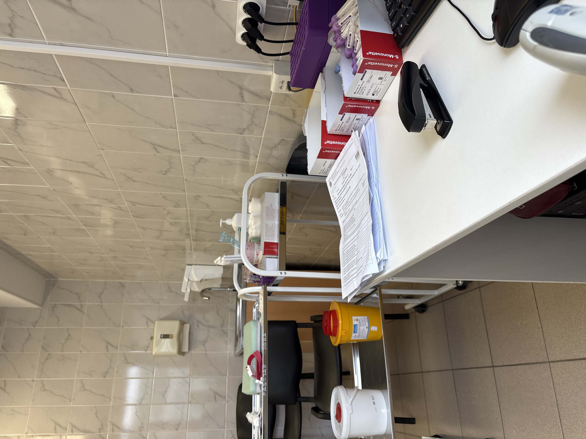 The image shows a corner of what appears to be a medical or laboratory setting. A white table occupies the right side of the frame, holding items like a keyboard, mouse, stapler, and boxes of "S-Monovette" blood collection tubes. A metal cart with wheels is positioned next to the table, containing various supplies including liquid soaps, blood collection containers, and sharps containers. The background features tiled walls in a neutral tone, typical of a clinical environment.
