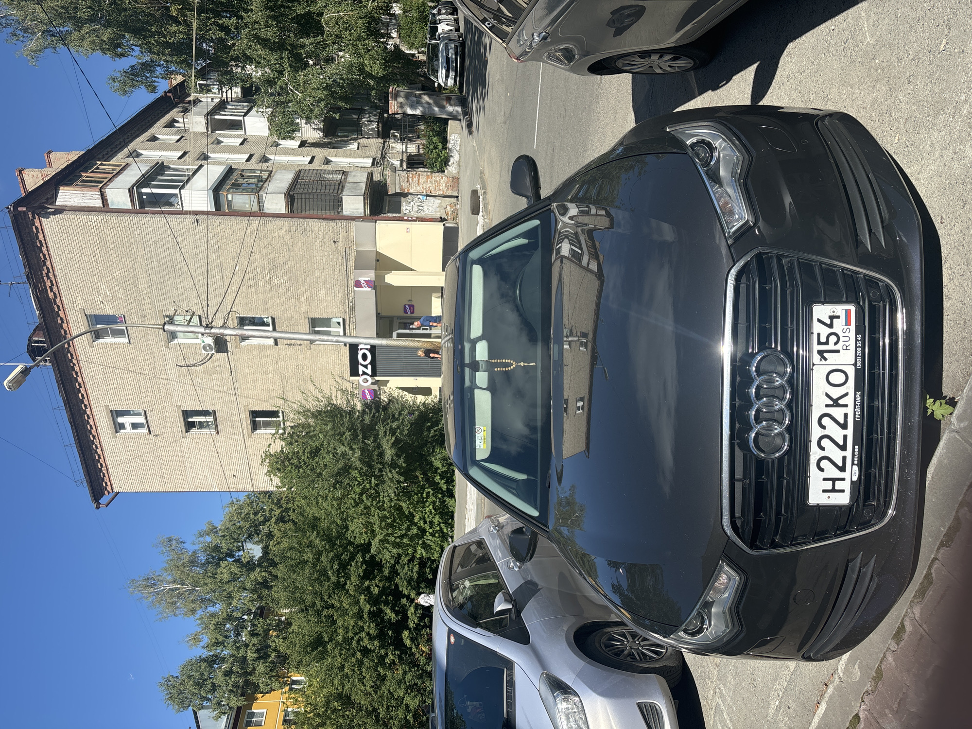 A dark grey Audi A6 sedan is parked on a paved surface, facing the camera. The car's license plate reads "H222KO 154 RUS". To the left of the Audi, a silver hatchback is parked. In the background, a light brick building with multiple floors and balconies is visible. A store with "OZON" signage is on the ground floor of the building. Trees and a clear blue sky complete the scene. The location is identified as Novosibirsk, Russia.