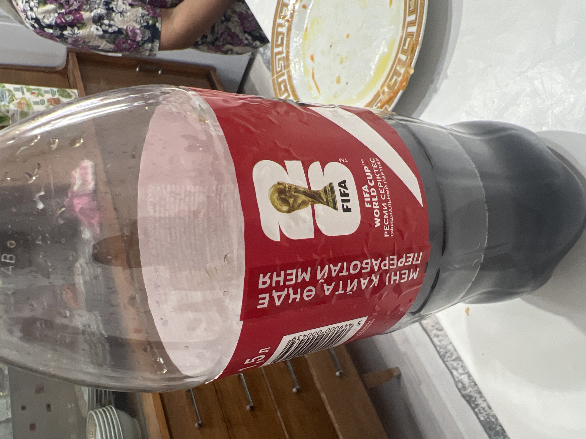 A partially filled Coca-Cola bottle, featuring a red label with FIFA World Cup branding and text in Cyrillic, stands on a white table. The label displays the FIFA World Cup trophy. To the left of the bottle, a wooden dresser with drawers is visible. In the background, a person wearing a floral patterned shirt is partially visible, along with a shelf unit containing various items. A used plate with remnants of food is situated to the right of the bottle. The setting appears to be an indoor domestic environment.