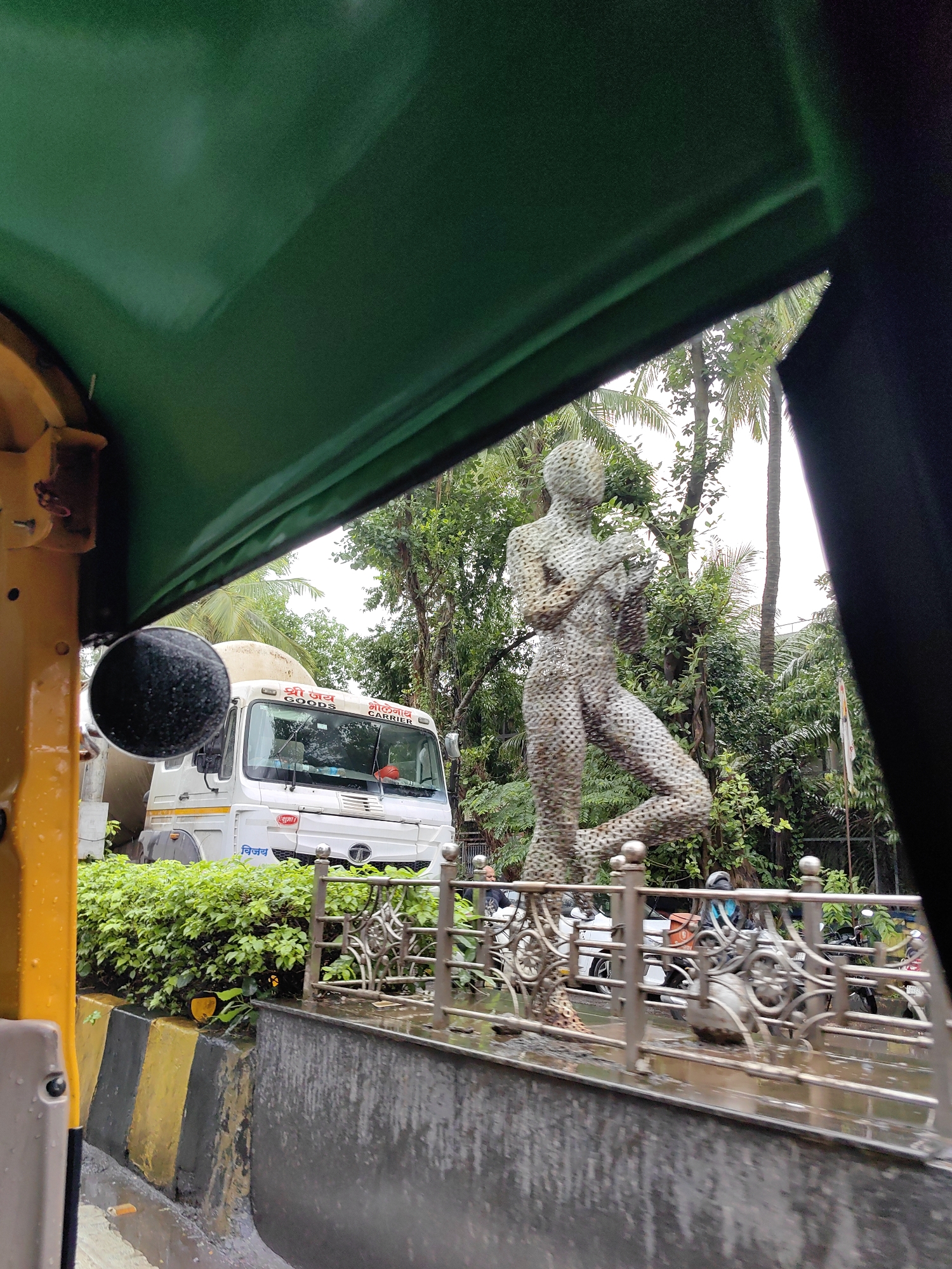An auto-rickshaw's green canopy is in the foreground, obscuring the top portion of the image. To the right, a black interior panel is visible. Below the canopy, on the left, a yellow portion of the auto-rickshaw is visible, along with a circular black object, possibly a side mirror.
In the middle ground, a white truck with a cement mixer drum is parked. The truck has Hindi text on it, including "श्री जय" and "GOODS". A statue of a human figure, made of wire mesh, stands to the right of the truck. The statue is in a dynamic pose, with one leg raised.
Behind the truck and statue, lush green trees and foliage are visible, indicating a tropical environment. A flagpole is visible on the far right.
In the foreground, a wet road surface is visible, with yellow and black striped barriers along the side. A metal railing with an ornate design runs in front of the truck and statue, separating them from the road. Several motorcycles and a white car are visible behind the railing. The overall scene suggests an outdoor setting in a city, possibly on a cloudy or rainy day, given the wet surfaces.
The scene is located in Mumbai, India, specifically in the Bandra area, where many contemporary sculptures and public art installations are found. The presence of an auto-rickshaw, the Hindi text on the truck, and the tropical vegetation are all consistent with this location.