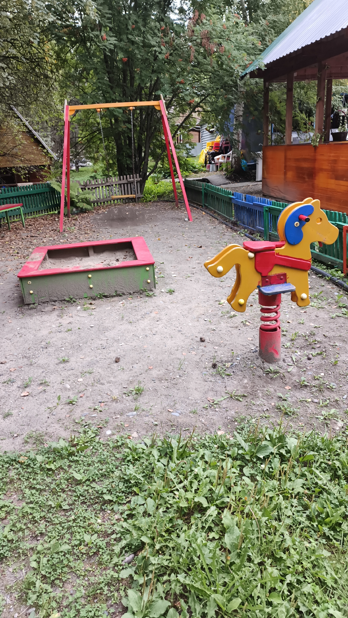 This is a photo of a playground in Katun', Russia. The playground features a red and yellow swing set, a sandbox with a green and red frame, and a yellow rocking horse. The ground is covered in dirt and some grass. In the background, there is a wooden structure with a green roof, possibly a gazebo or pavilion, and trees with red berries. The scene is outdoors, under a cloudy sky. There are no people visible in the image.