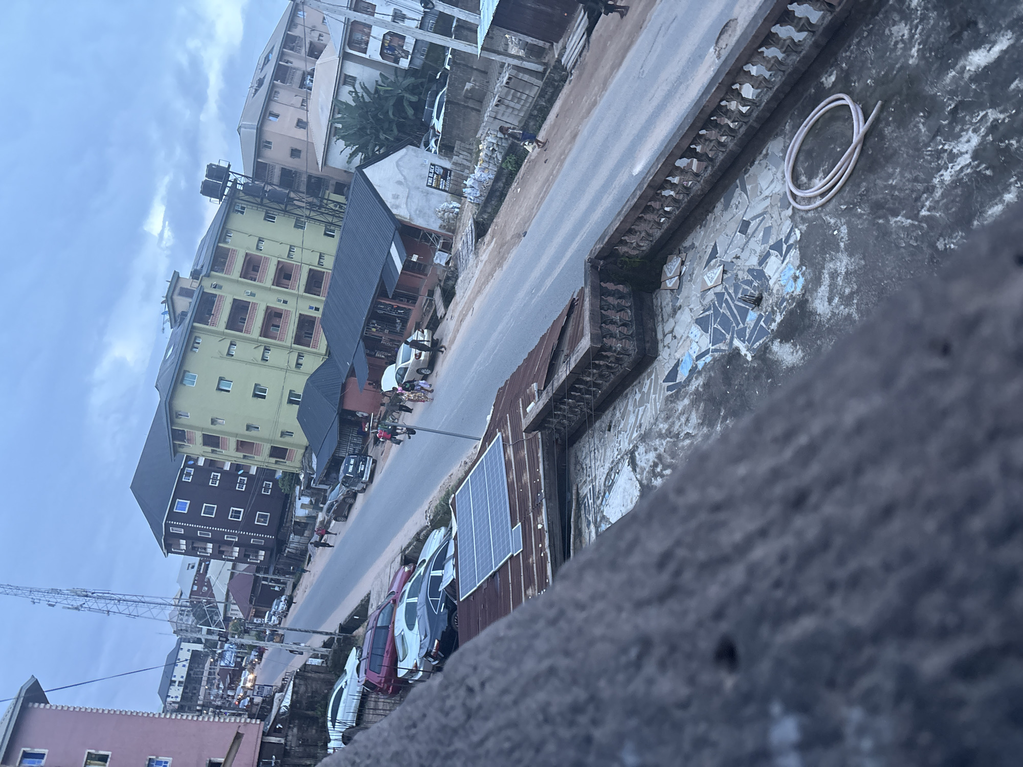 This is a high-angle, full shot of a street scene in Nkpor, Nigeria, under an overcast sky. The image focuses on a street lined with multi-story buildings and parked vehicles. In the foreground, a textured concrete balcony with a decorative railing overlooks the street. On the balcony, a coiled hose lies on the ground, and a solar panel array is installed on a corrugated metal roof.
The street is paved and shows signs of wear. Several cars are parked along the side of the road, including a white sedan, a dark SUV, and a black sedan. Pedestrians are visible on the street and sidewalks. Some are walking, while others appear to be standing or interacting. The buildings vary in color and architectural style, with some featuring balconies and windows. One prominent building is painted lime green, another is dark brown, and a third is pale pink. A tall cellular tower is visible in the distance, behind the buildings. The overall impression is of a bustling urban environment.