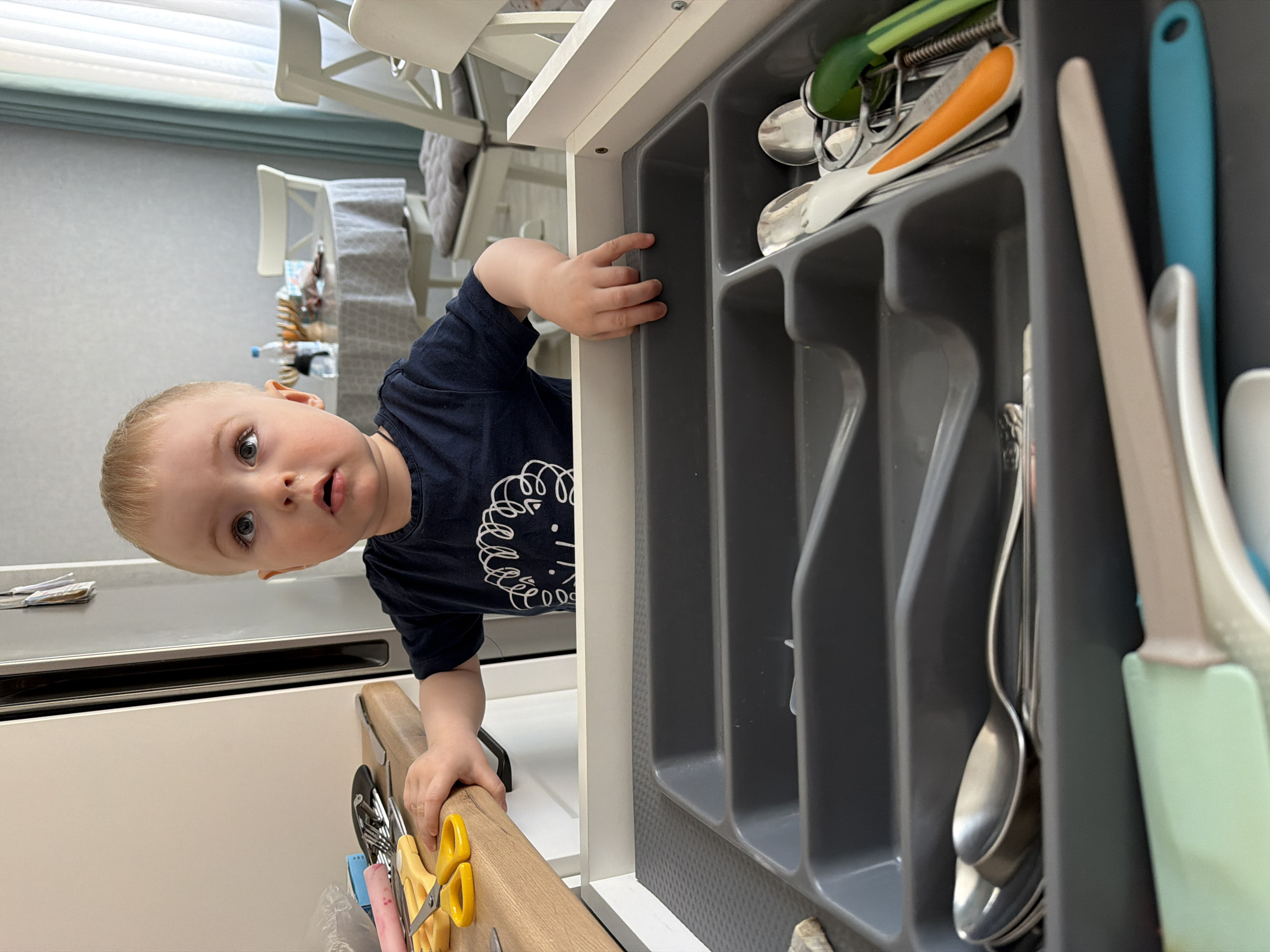 A child is standing next to an open drawer of a kitchen cabinet. The drawer contains a gray utensil organizer with various kitchen utensils, including spatulas, spoons, and scissors. The child is wearing a dark blue T-shirt with a white graphic on it. The kitchen has white cabinets and is lit by daylight. The image does not provide evidence of a location in Votkinsk, Russia.