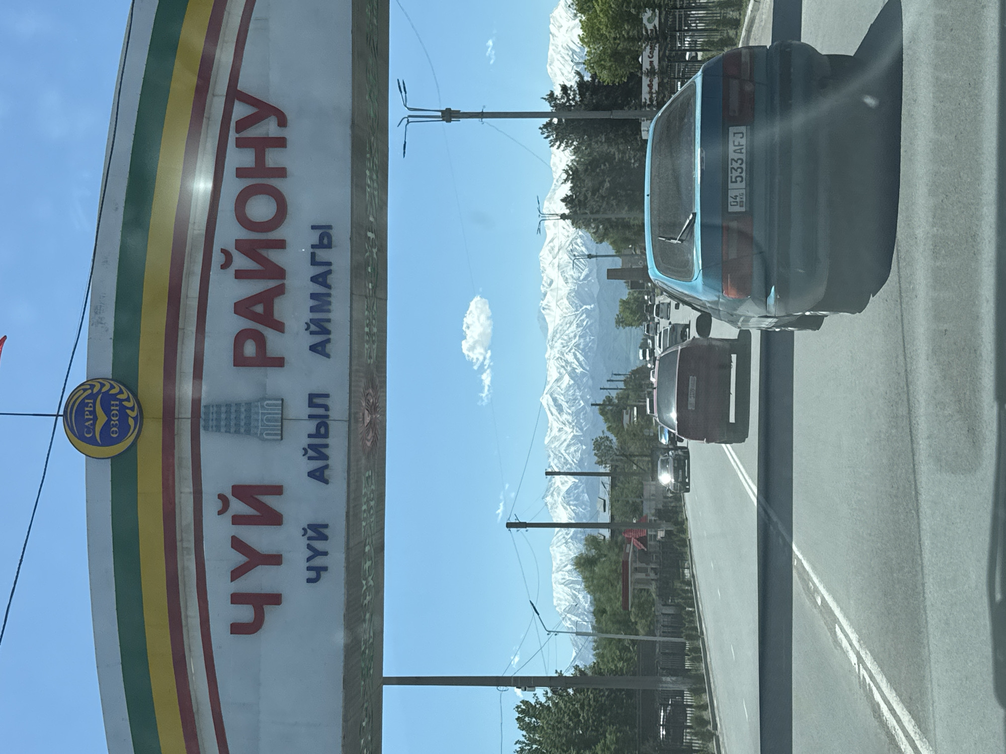 The image shows a street scene in Tokmok City, Kyrgyzstan, framed by a road sign and vehicle traffic. The road sign on the left features the text "ЧҮЙ РАЙОНУ" and "ЧҮЙ АЙЫЛ АЙМАГЫ," indicating the location is in the Chüy region. A registration plate number 04 533 AFJ on a blue car in the foreground suggests the vehicle is registered in the same region, as 04 is the regional code for Chüy Oblast. A line of cars is visible on the road ahead. Snowy mountain ranges are visible in the background, under a blue sky with scattered clouds. Overhead power lines and street lamps are present along the roadside.
