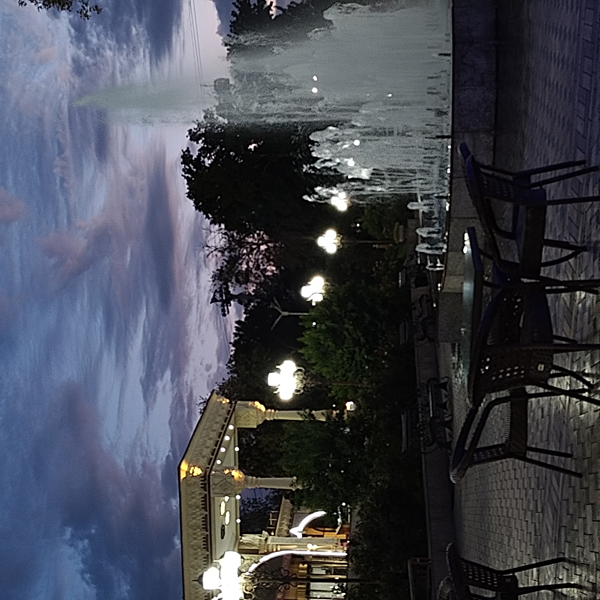 The image depicts an outdoor park scene at dusk in Khujand, Tajikistan. In the foreground, several dark wicker chairs are arranged around tables on a cobblestone patio. To the right, a large, multi-tiered fountain is actively spraying water. In the background, a white, ornate gazebo-like structure is illuminated by decorative lights. Trees and lampposts are visible throughout the scene. The sky is a deep twilight blue with dramatic clouds. The overall impression is of a serene evening in a public recreational area.