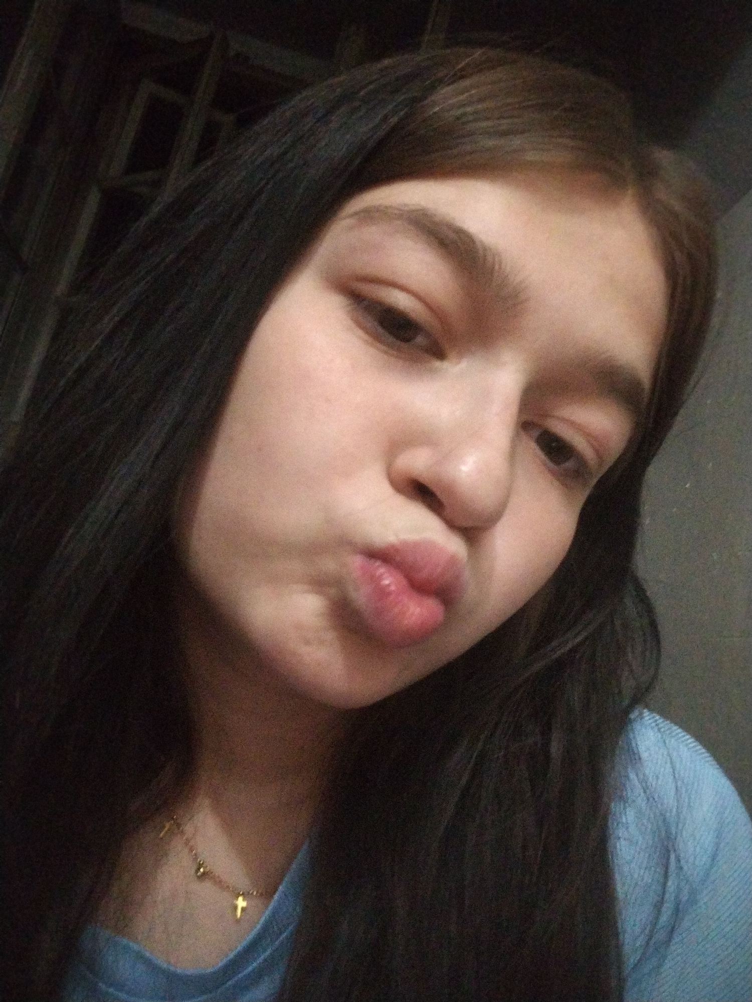 A young person with dark hair and fair skin is taking a selfie. They are wearing a light blue collared shirt and a delicate gold necklace with small cross pendants. Their lips are pursed, and they are looking towards the camera. The background appears to be an indoor setting with a dark, textured wall and some indistinct objects in the upper left corner. The lighting is dim, suggesting it might be evening or an indoor space with limited light.