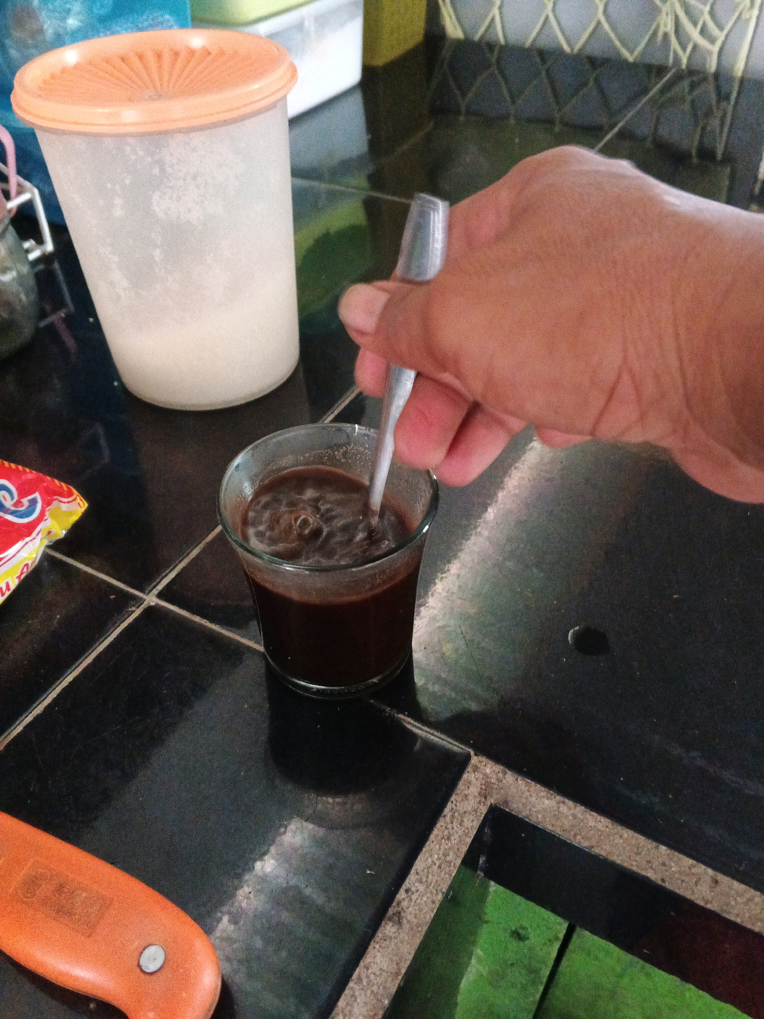 In the city of Brangan Mulya, Indonesia, a close-up shot captures a hand stirring a dark liquid in a small glass, presumably coffee, with a metal spoon. The hand, with visible skin texture, is holding the spoon, creating ripples in the beverage. Adjacent to the glass, a brightly colored packet, possibly of instant coffee or a related product, rests on a black tiled surface. In the background, a translucent white container with an orange lid is partially visible, suggesting it might hold sugar or creamer. The overall environment appears to be a domestic or casual setting, indicated by the tiled countertop and the presence of food-related items.