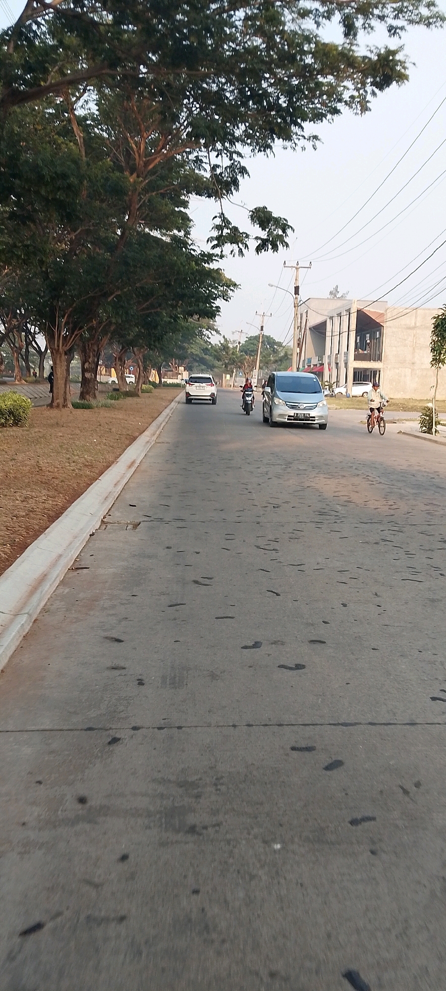 A street scene in Satriajaya, Indonesia, captures a grey asphalt road lined with mature trees on the left. The road is partially wet with dark patches, suggesting recent rain or spills. Several vehicles are visible: a white SUV is driving away from the camera, followed by a motorcycle with a rider wearing a red shirt. A silver minivan is moving towards the camera, with a driver visible. Further down the road, a person rides a bicycle. On the right side of the road, modern buildings with large windows are under construction or recently completed. Power lines are strung across the sky, supported by utility poles. The overall lighting suggests it is daytime, possibly late afternoon, with a clear, pale sky.