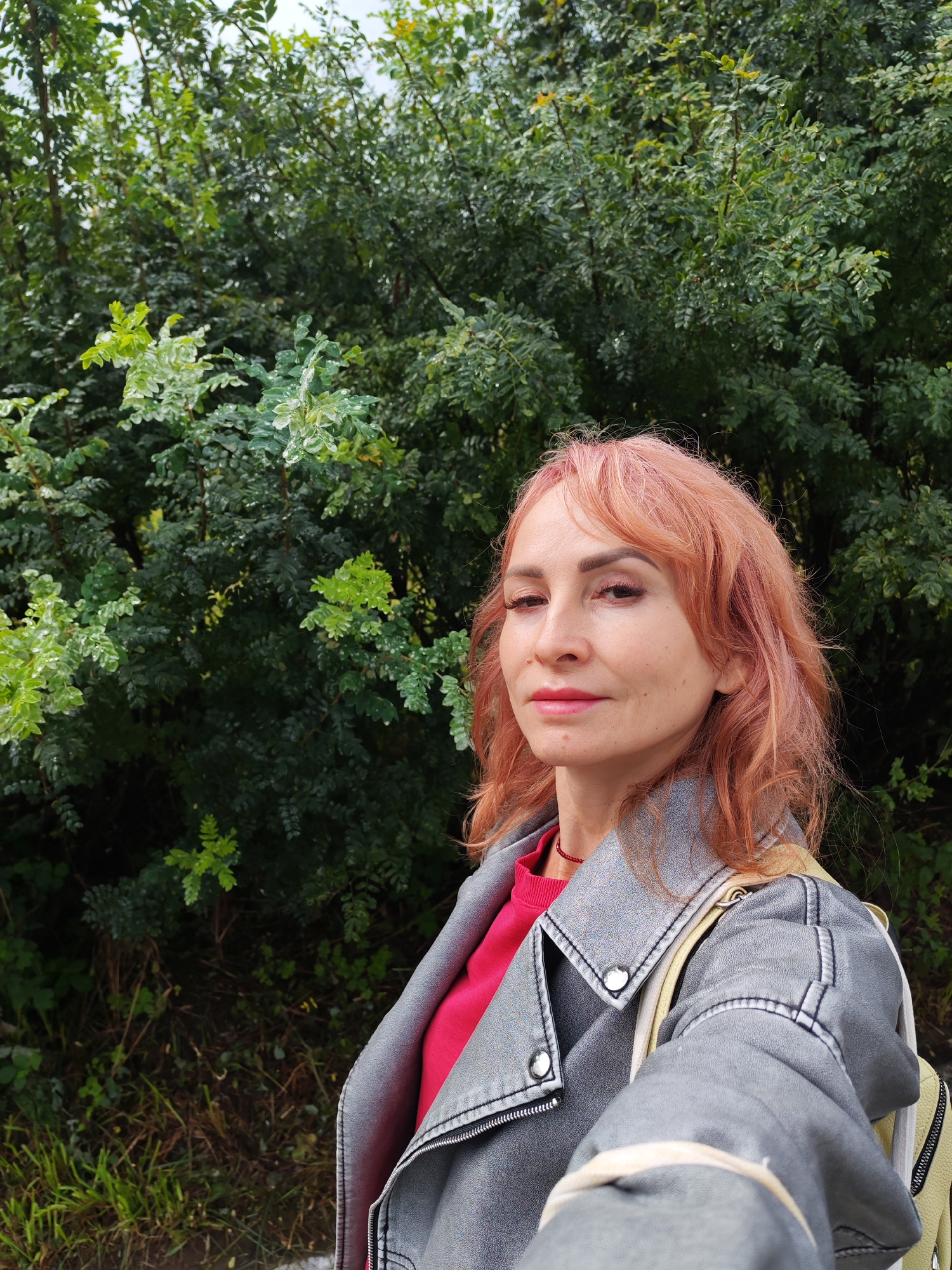 A woman with pink hair, wearing a gray jacket over a pink shirt, poses for a selfie in front of dense green foliage. The leaves are small and numerous, with some appearing lighter green, possibly due to recent rain as water droplets are visible on some of them. The woman's makeup includes eye shadow and lip gloss. She has a red necklace on. A yellow backpack strap is visible over her shoulder. The lighting suggests an outdoor environment, possibly a park or a wooded area. The background is a wall of green leaves and branches, with some areas of darker shadow and lighter patches indicating dappled sunlight. The overall impression is of a casual outdoor portrait.