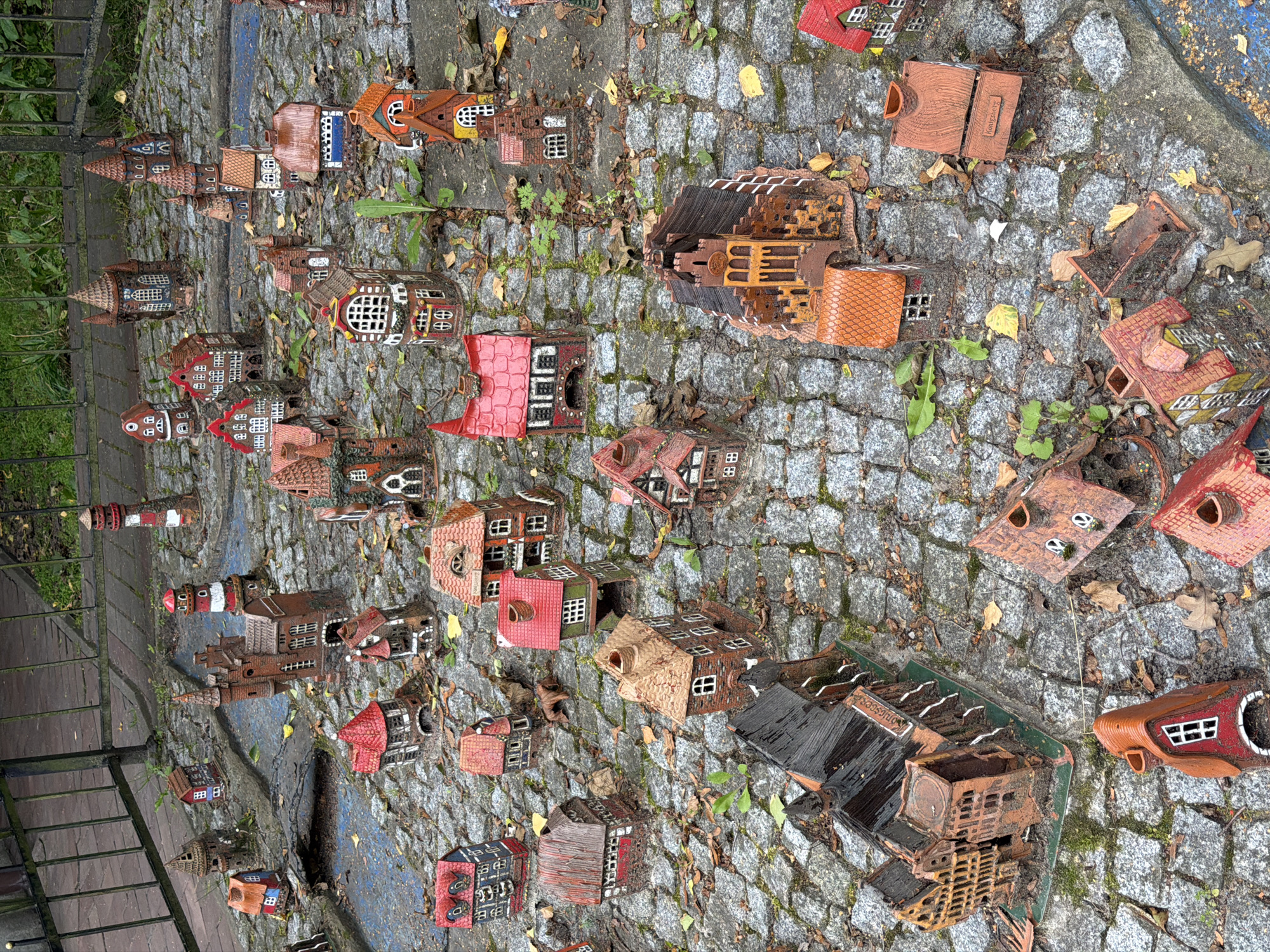The image shows a miniature village scene set on a cobblestone ground, located in the city of Svetlogorsk, Russia. The village consists of numerous small, intricately designed ceramic houses, reminiscent of old European architecture. The houses vary in size, shape, and color, with many featuring red roofs, brick-like facades, and detailed windows and doors. Some structures resemble townhouses, while others appear to be churches or other public buildings, and two lighthouses are also visible. The scene is outdoors, with a metal fence and greenery visible in the background, suggesting a park or a public display area. Fallen leaves are scattered across the cobblestones and among the miniature houses, indicating an autumnal setting. No people are visible in the image.
