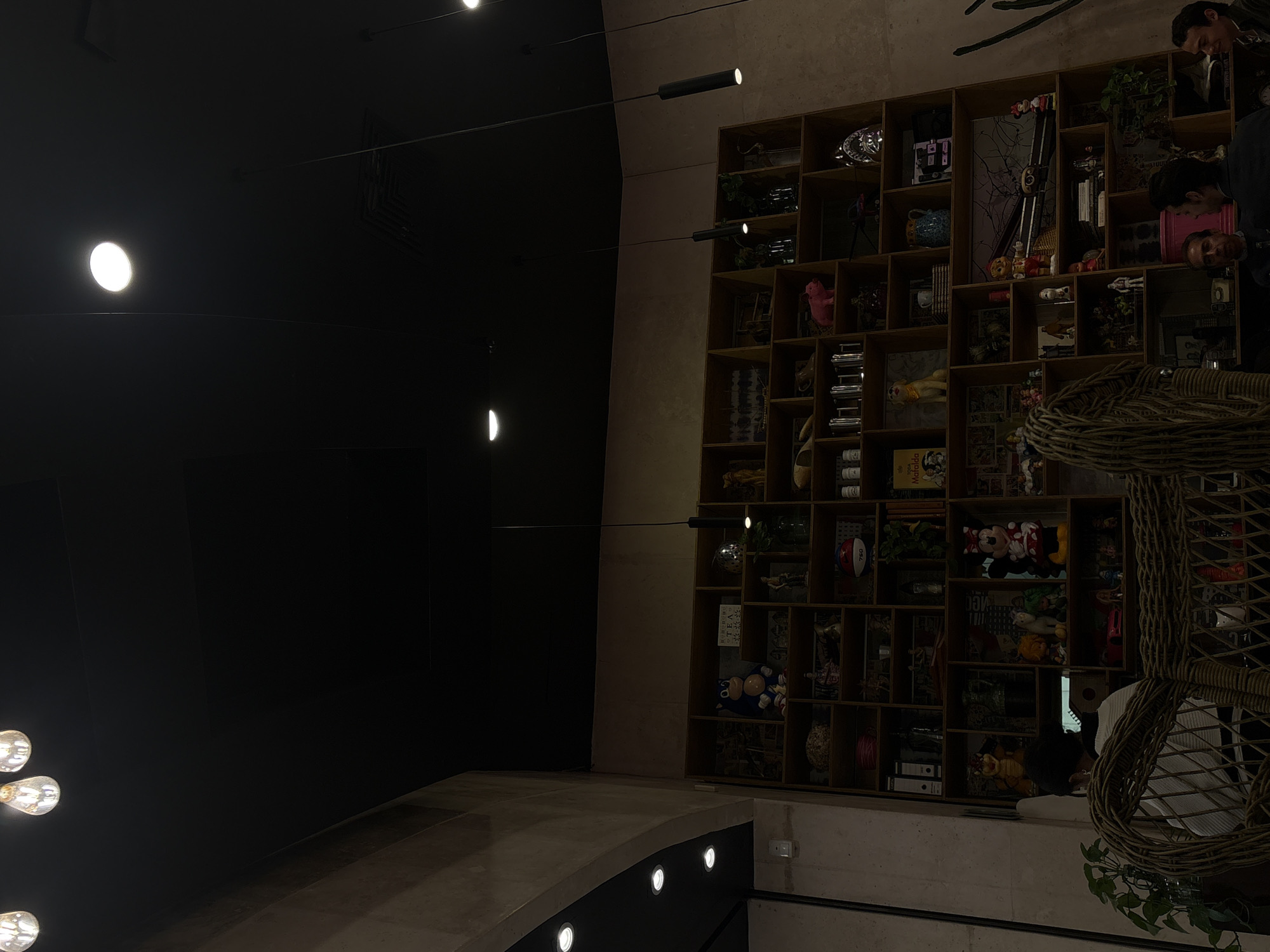 The image captures the interior of a space, likely a restaurant or lounge, located in the city of Puebla, Mexico. A large bookshelf is filled with eclectic items, including toys, books, plants, and decorative objects. To the left, a dark wall is punctuated by decorative light fixtures. Seating is visible, including a wicker chair. Several people are present in the image.