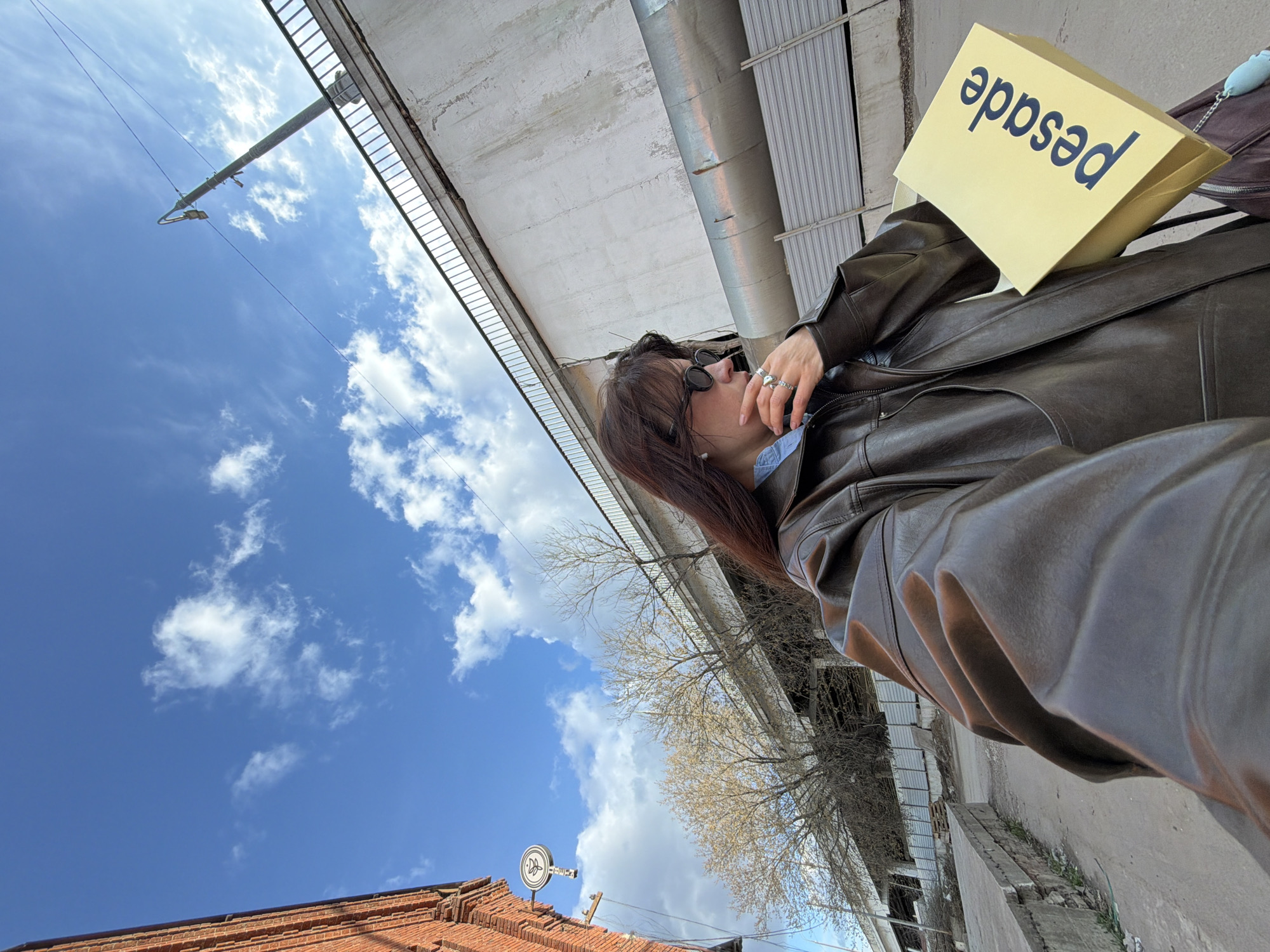 A woman in a brown leather jacket is standing under a bridge on a sunny day, holding a yellow shopping bag with the word "pesad" printed on it. The sky is blue with white clouds. In the background, there are trees and a red brick building. The location is Moscow, Russia.